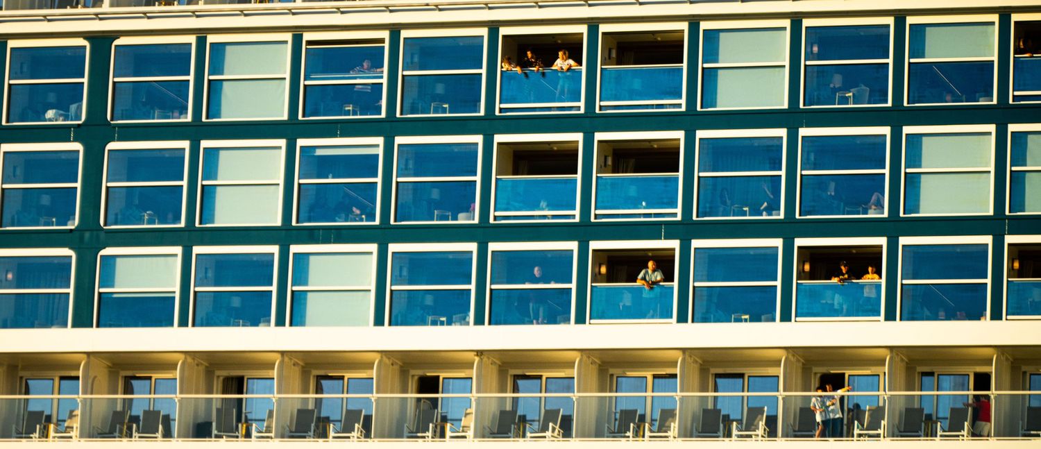Modern apartment building exterior with rows of blue-tinted windows and balconies reflecting bright sunlight.