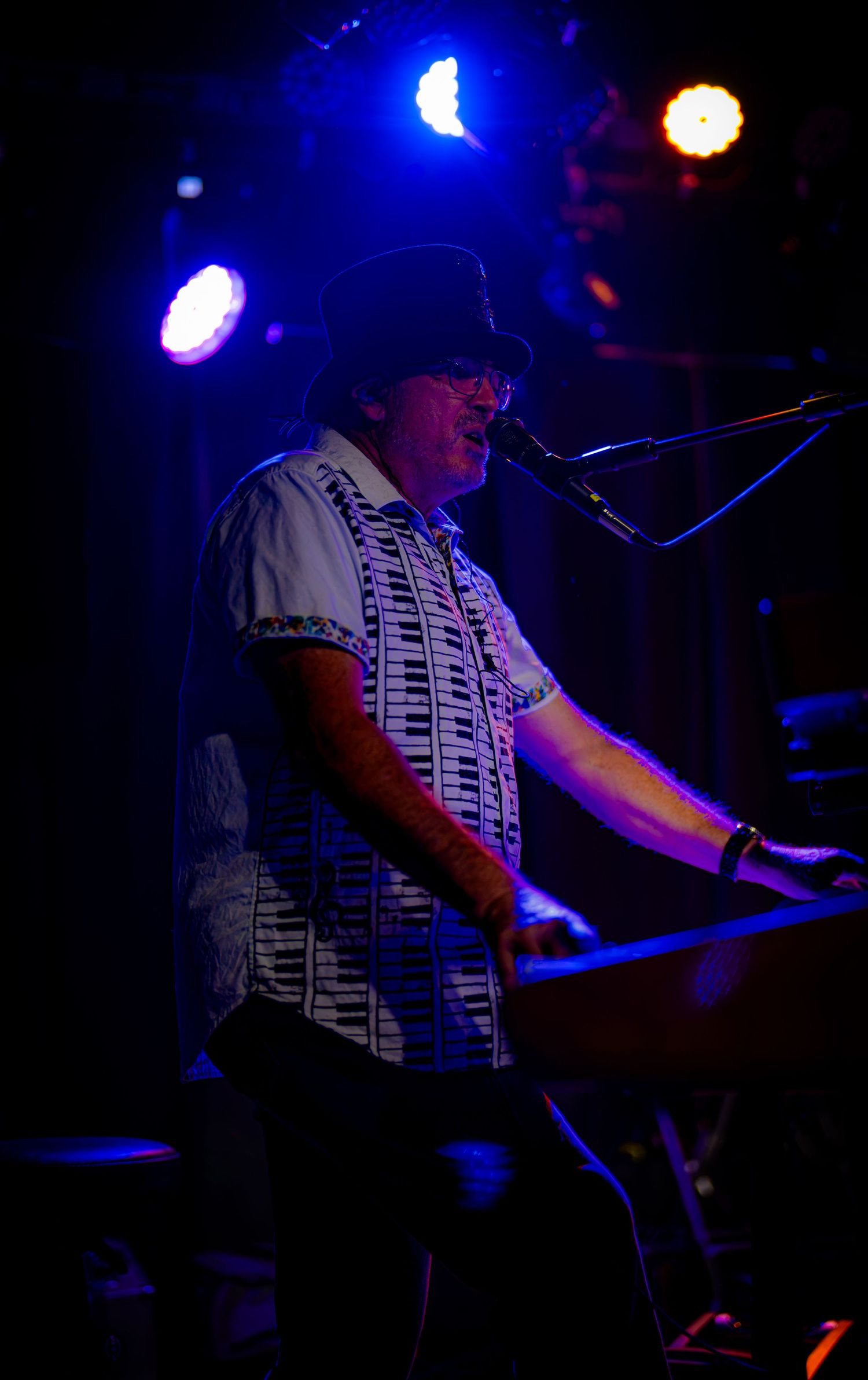 Musician playing keyboard on stage under blue and orange lights wearing a patterned shirt and bucket hat.