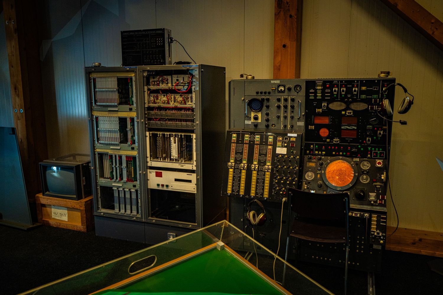 Vintage electronic control consoles with analog dials, switches and equipment racks in a wooden-walled room beside an old CRT monitor.
