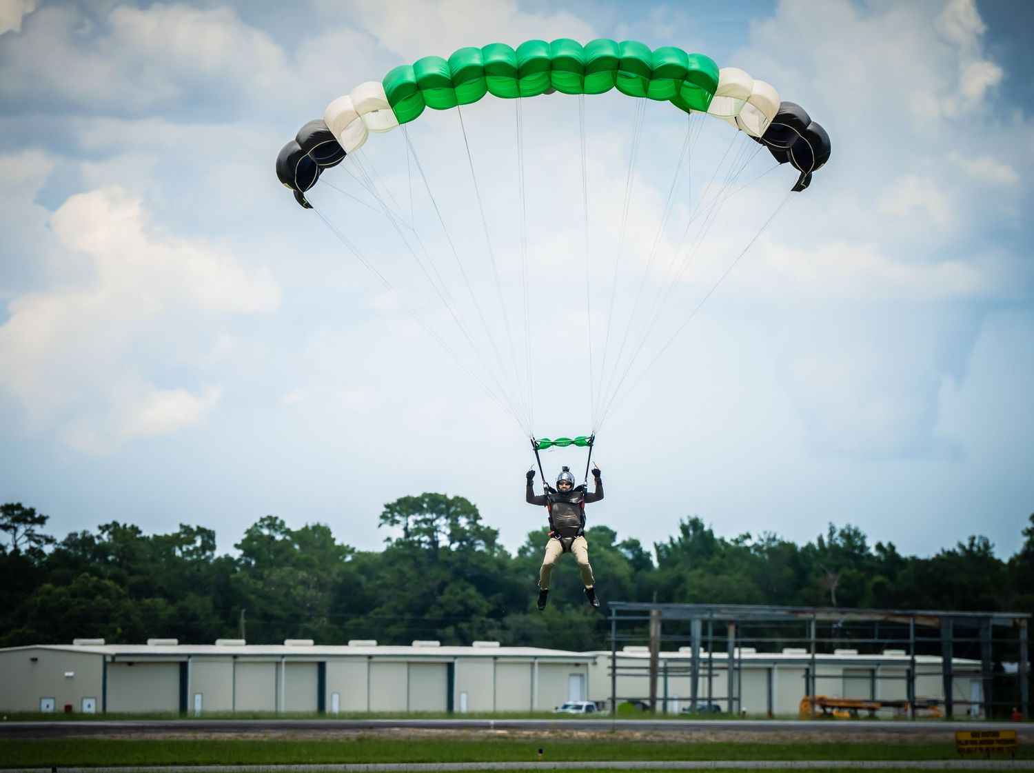 Bright green rectangular parachute gliding over an airfield with white buildings visible.