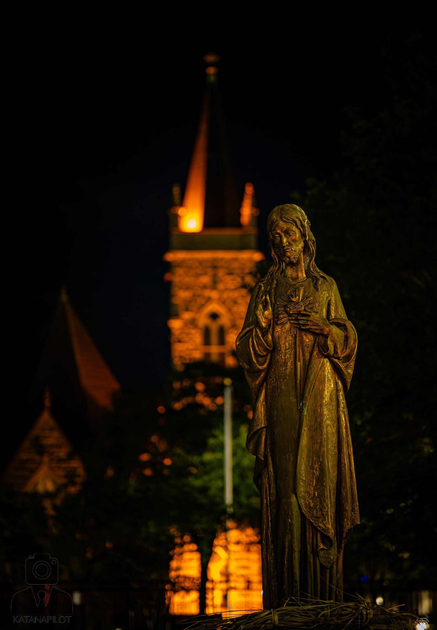 Bronze statue of Virgin Mary illuminated at night with glowing church steeple in background.
