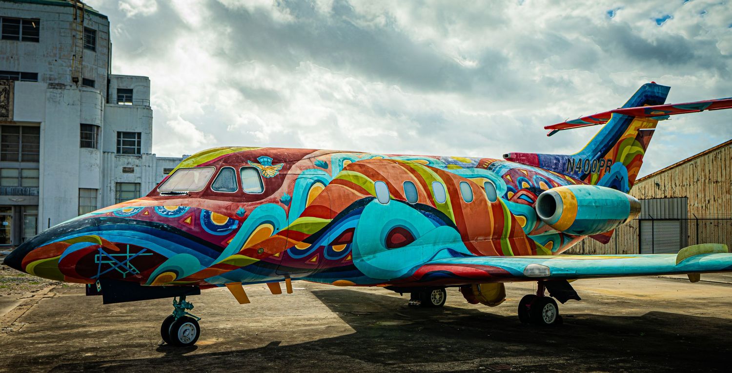 Colorful graffiti art painted small jet airplane parked on tarmac with vibrant abstract designs in blue, red, and yellow.