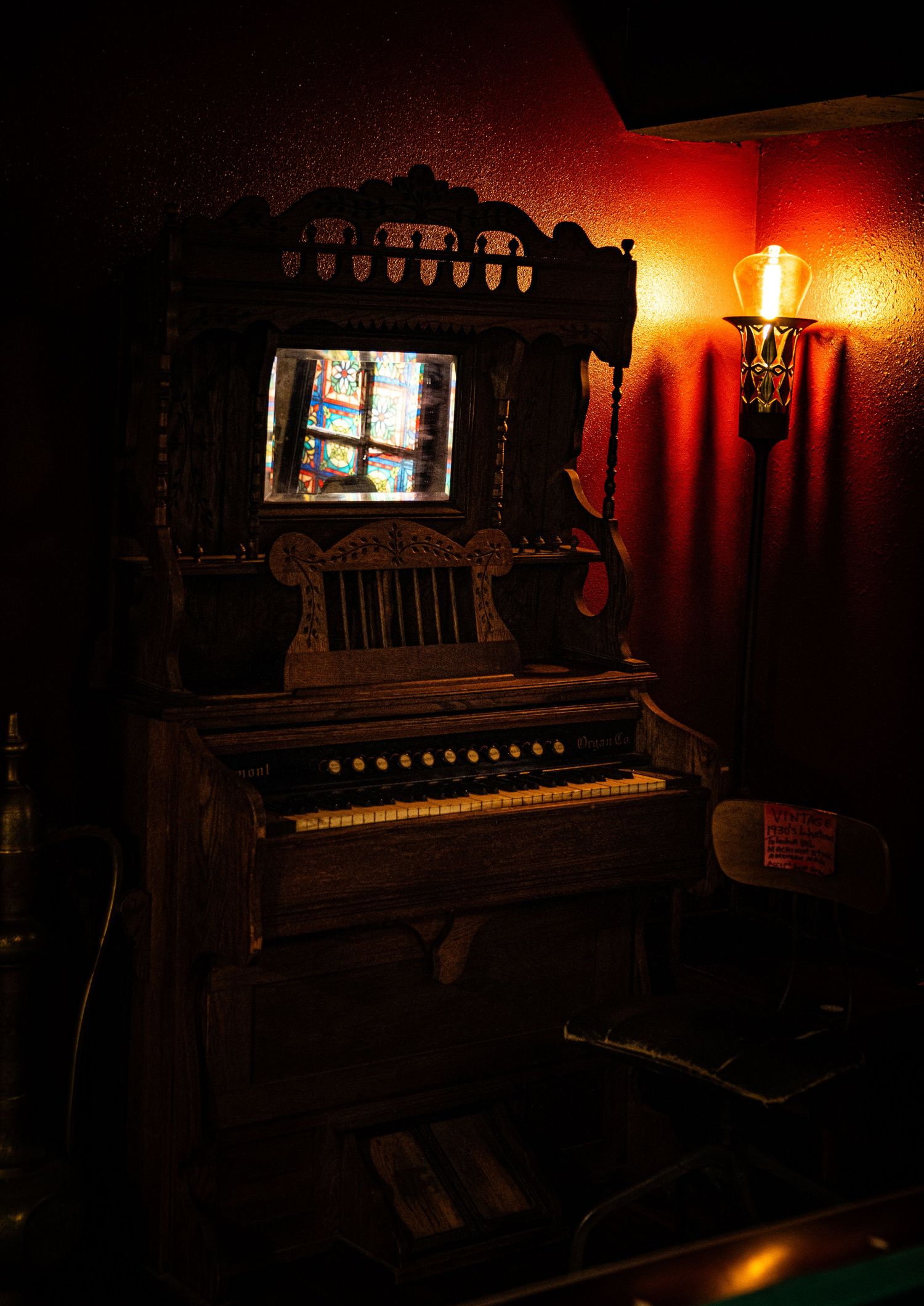 An antique pump organ sits in a dimly lit corner with warm ambient lighting from a wall sconce casting atmospheric glow.