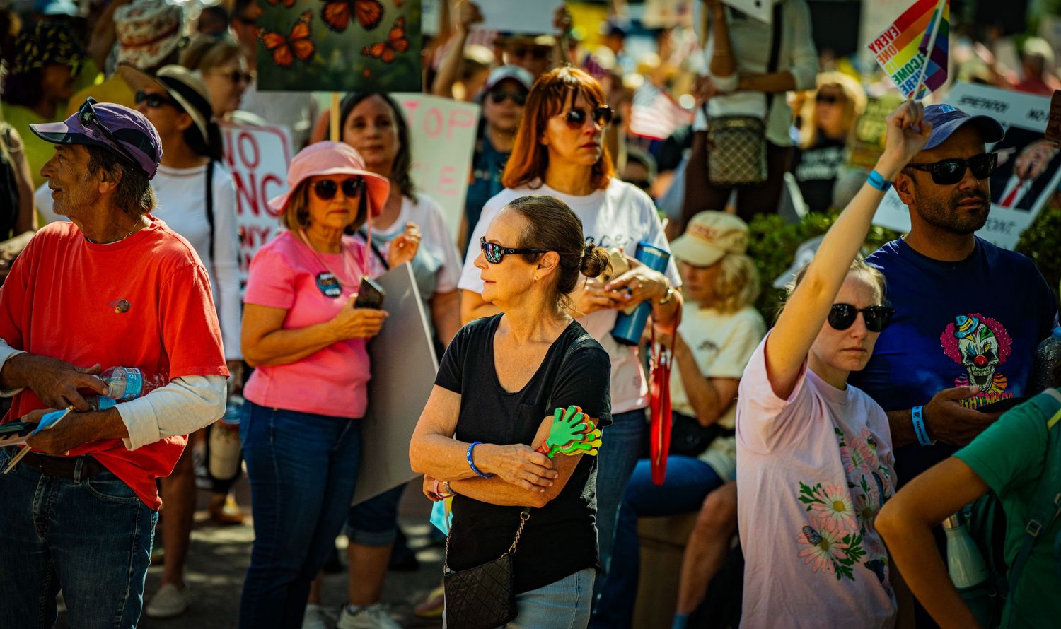 Demonstrators gather with signs and rainbow flags during a sunny protest event.