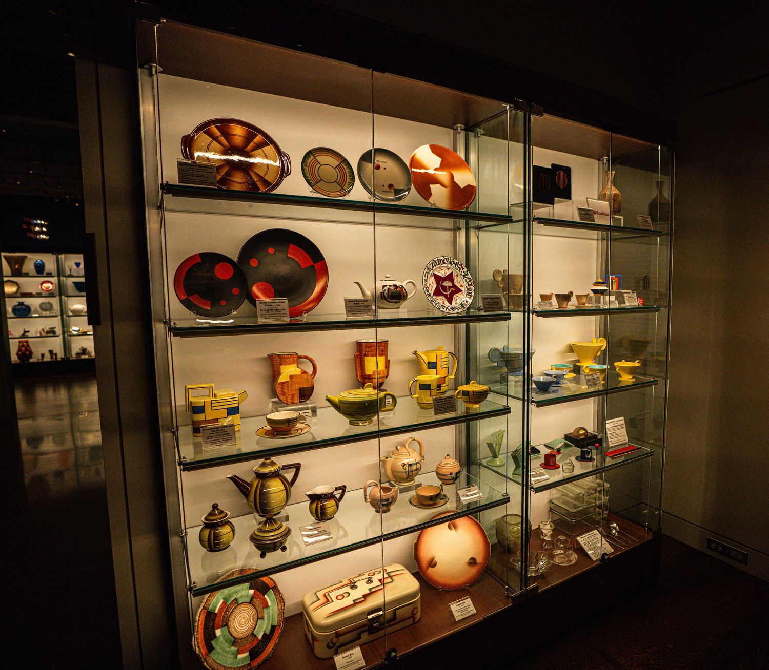 A glass display case illuminated in warm light showcases vintage decorative plates, cups, and collectible items on shelves.
