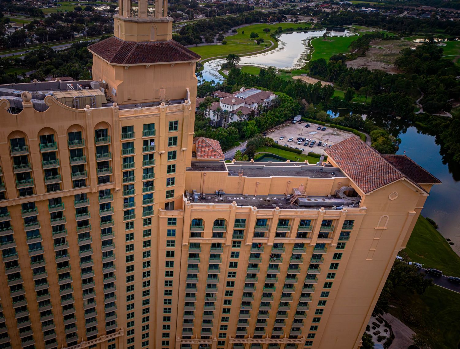 Aerial view of a large luxury hotel tower with terracotta roof, surrounded by lush greenery and a scenic lake.