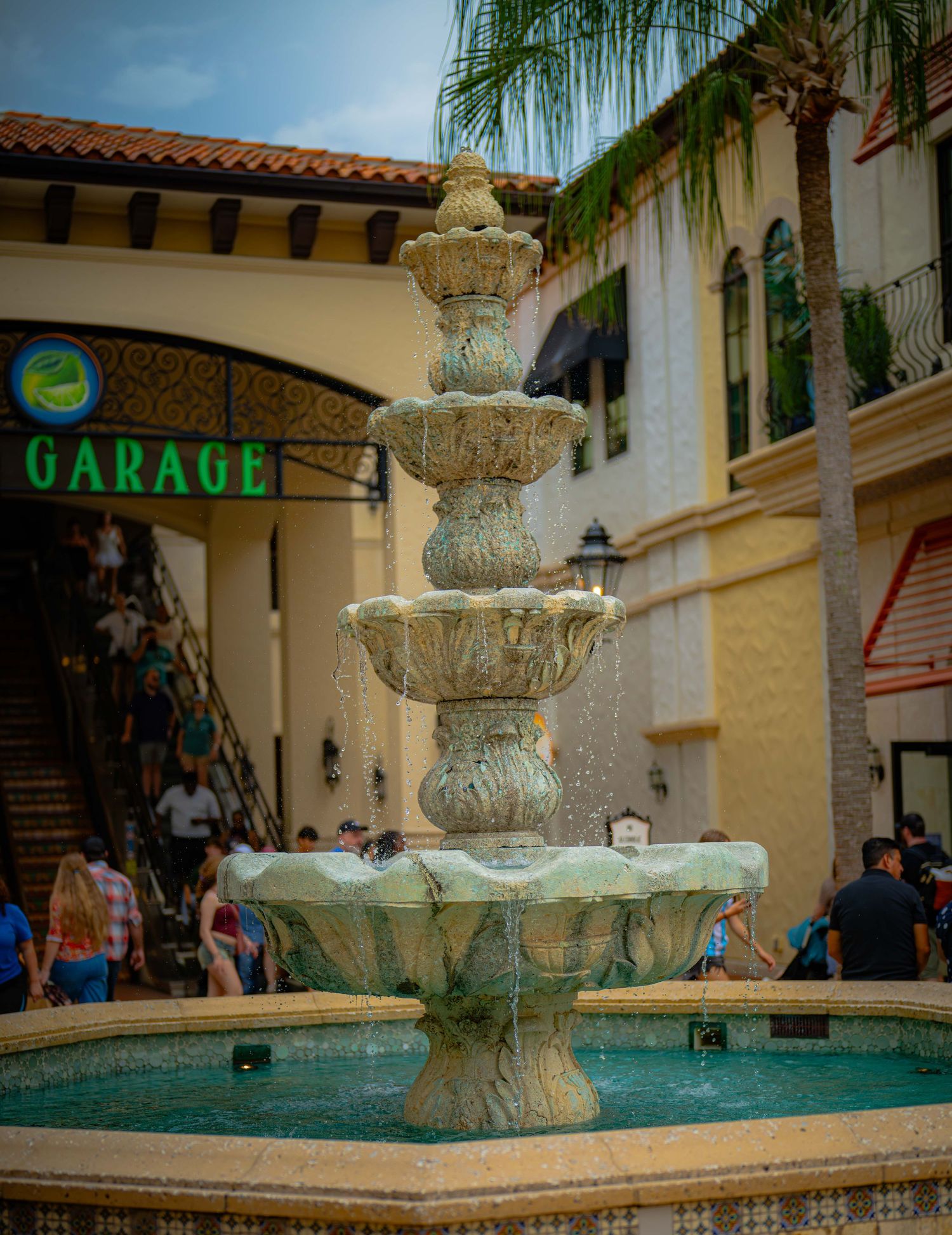 Multi-tiered stone fountain with cascading water in a bustling outdoor shopping plaza with tropical palms.