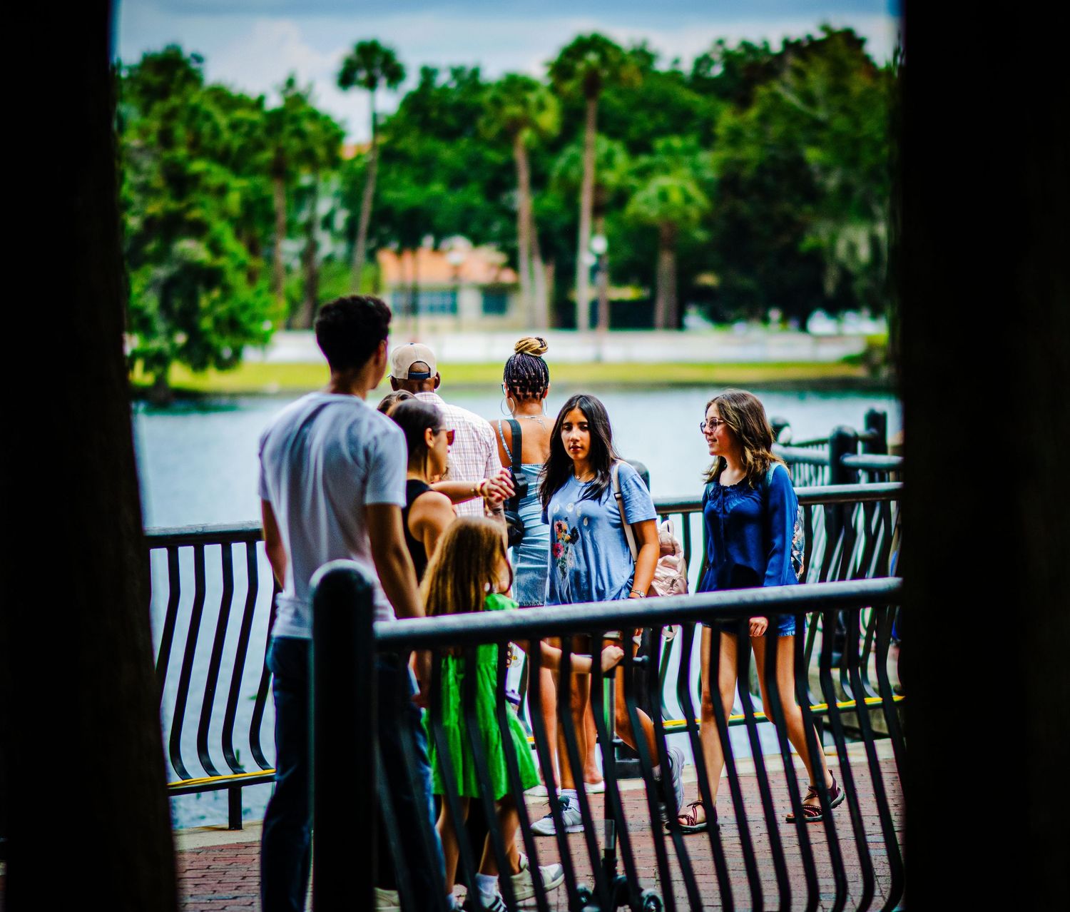 Group of teens socializing on a waterfront bridge surrounded by lush green trees on a sunny day.