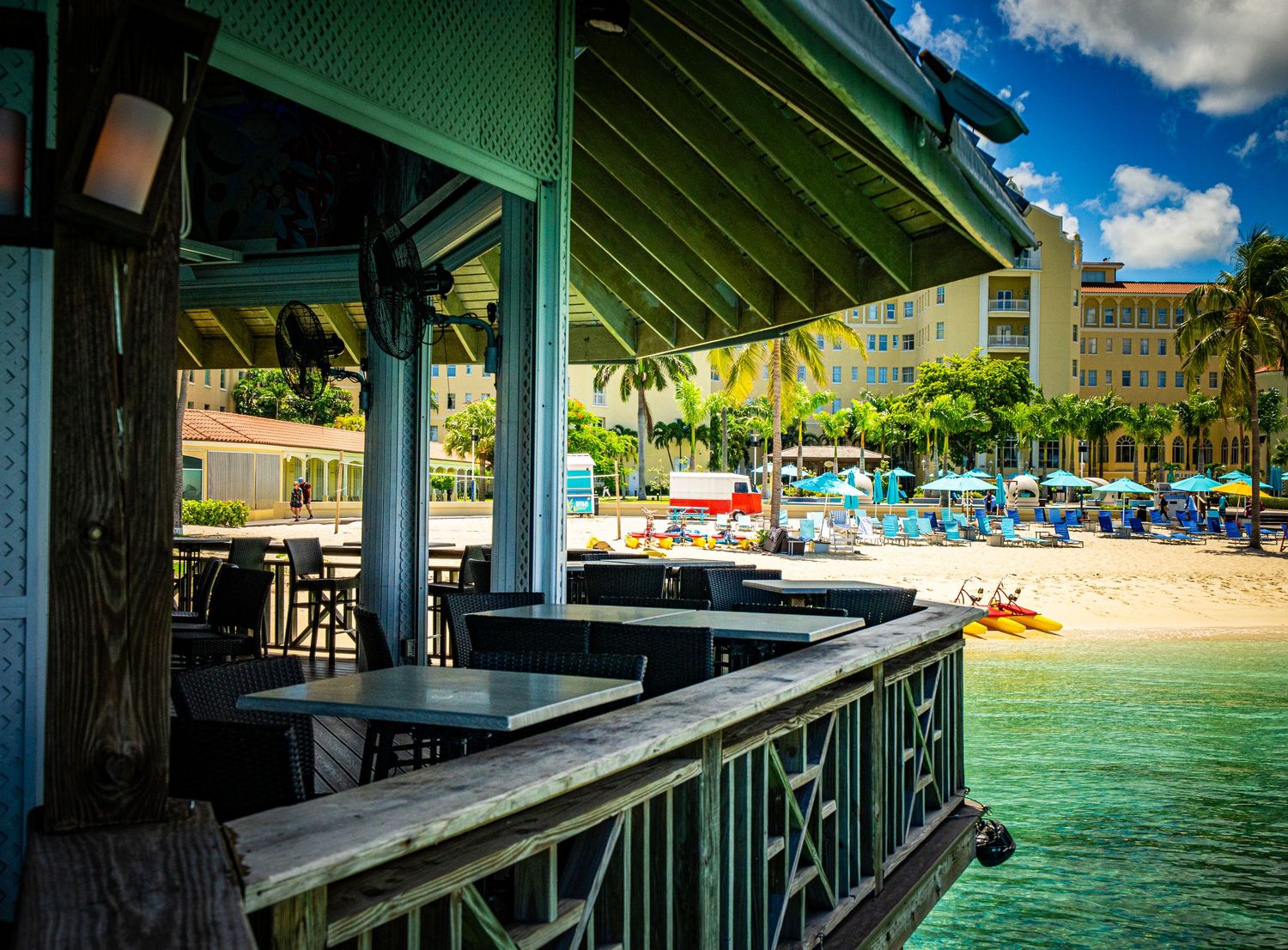 Tropical beachside bar with turquoise water, palm trees, and colorful resort in the background on a sunny day.