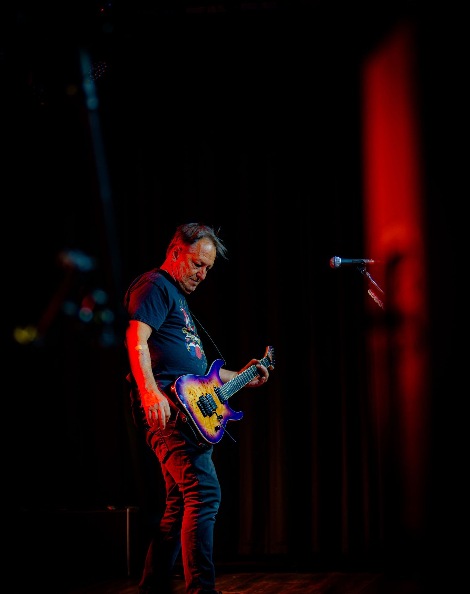 Male guitarist performing on dark stage with colorful guitar under red and blue stage lighting.
