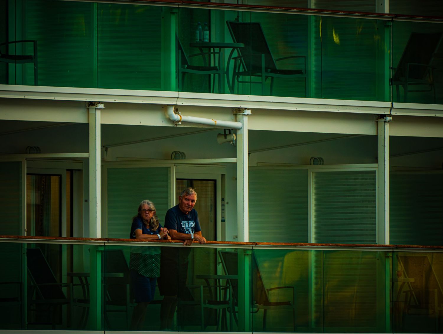 People observe from a balcony in a green-tinted building with glass windows and metal railings.