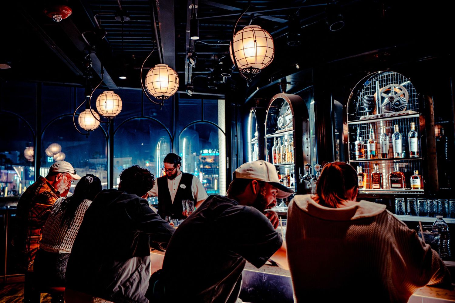 A dimly lit bar interior with glowing paper lanterns and patrons sitting at the counter during evening hours.