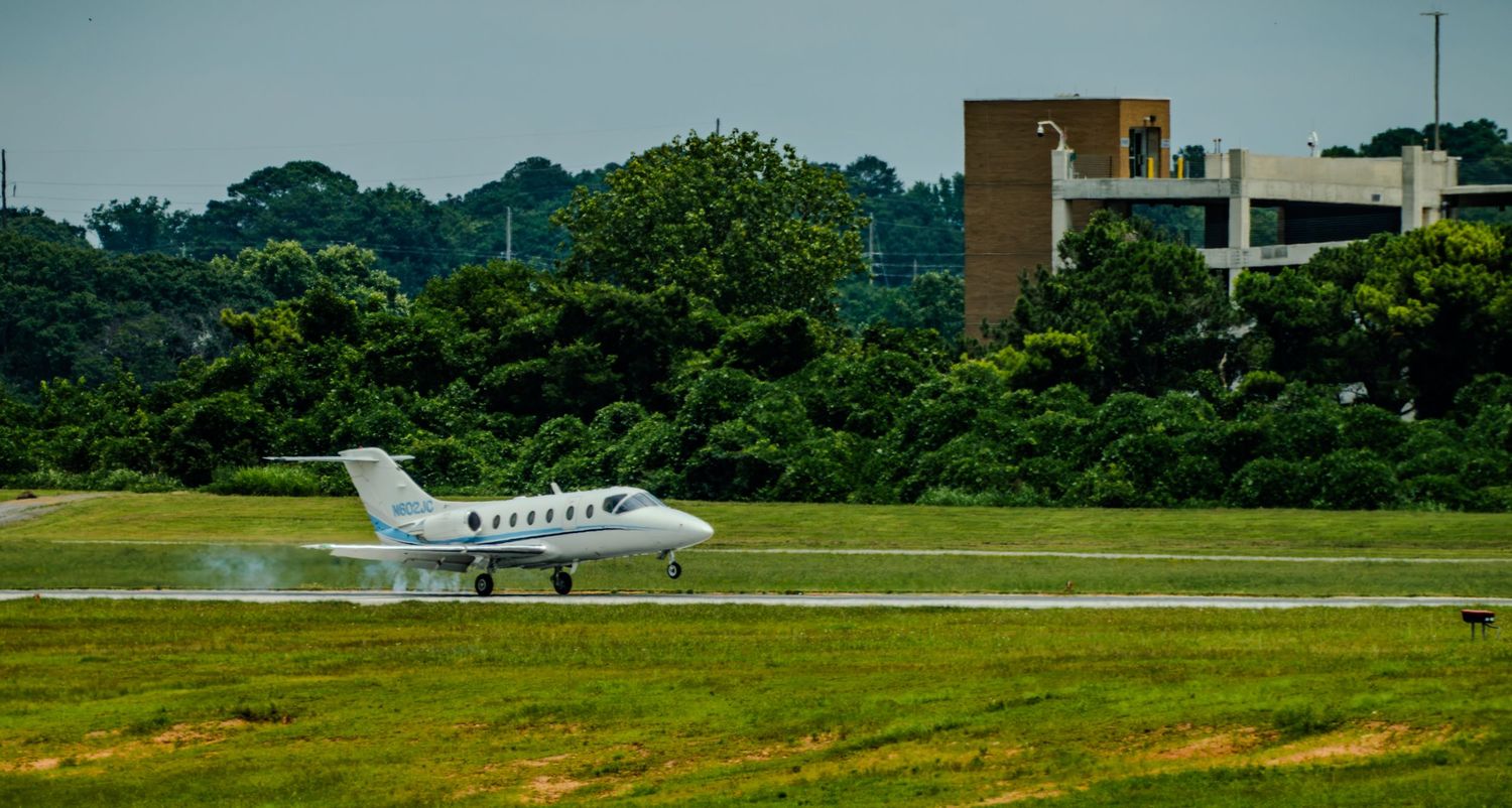 Small white private jet landing on a grass-lined runway with trees and a building in the background.
