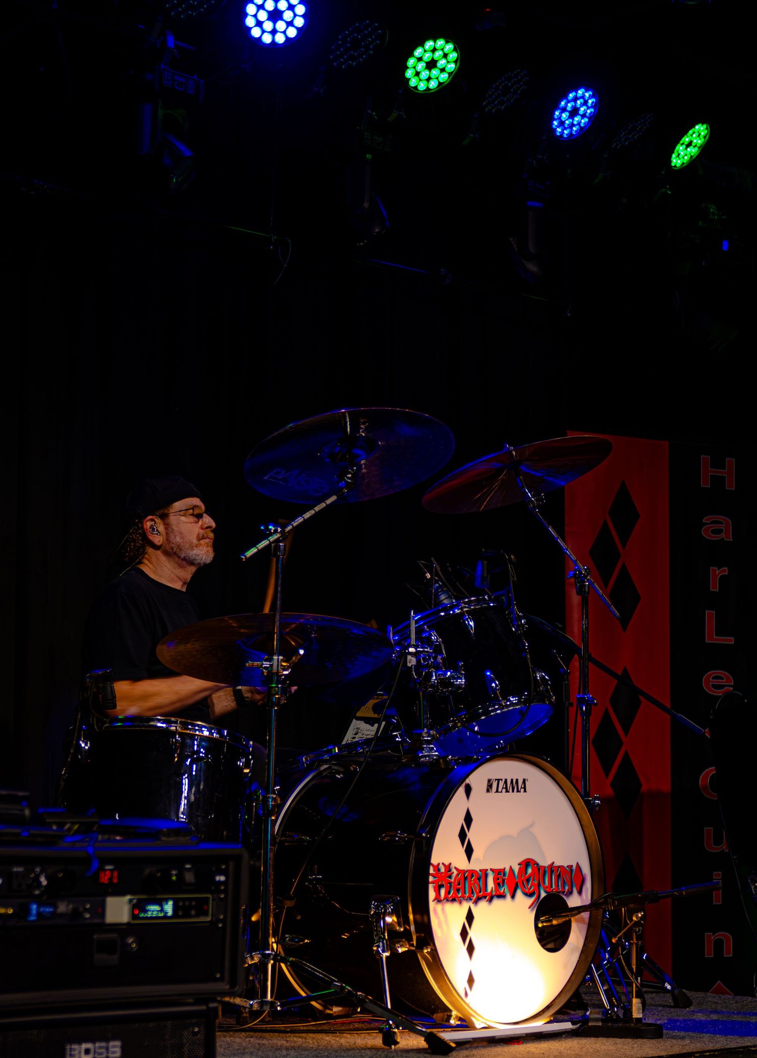 Drummer performing on stage with Tama drum kit featuring Harley Quinn branding under colorful stage lights.