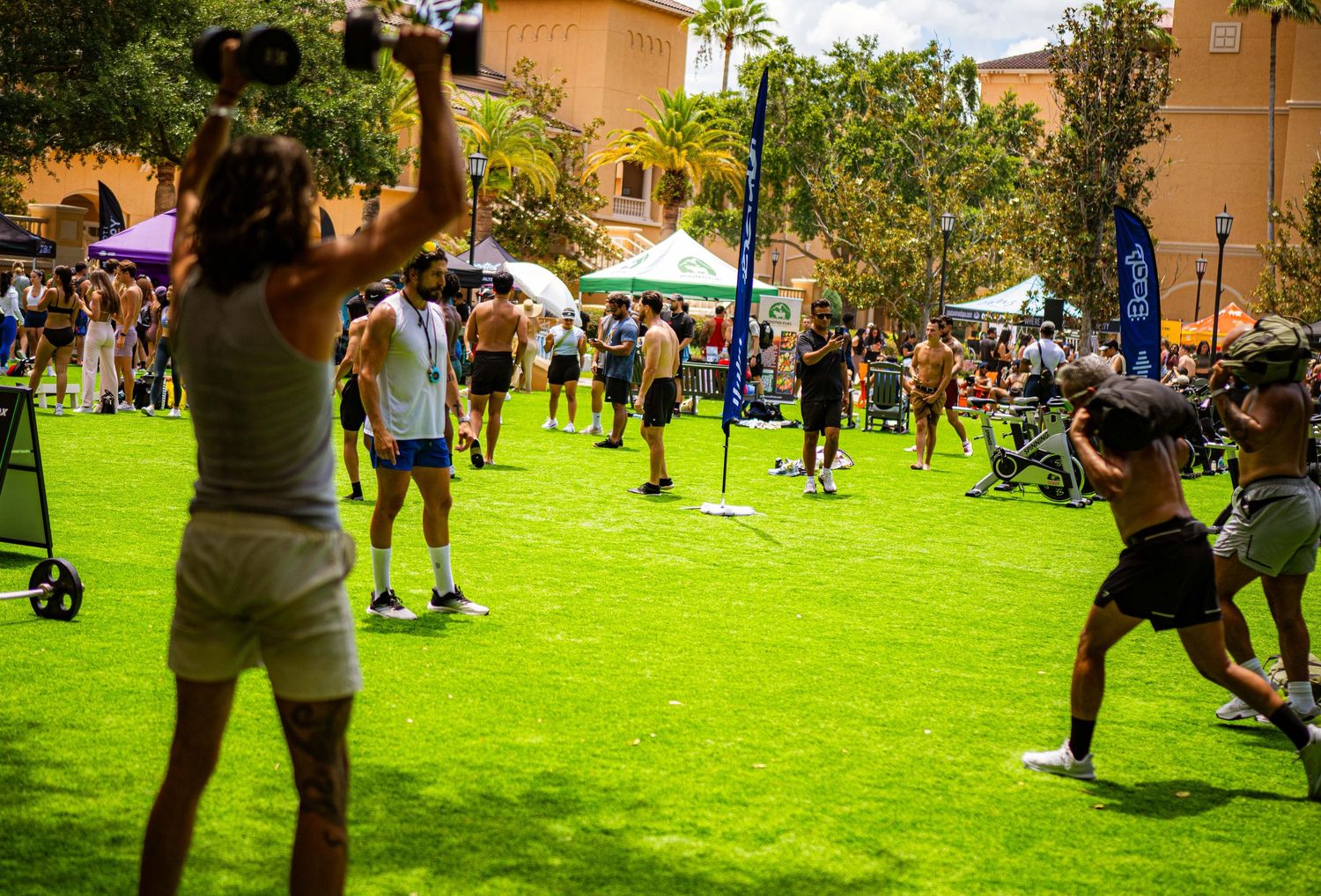 Outdoor fitness event on green grass with people exercising using dumbbells and equipment on a sunny day.