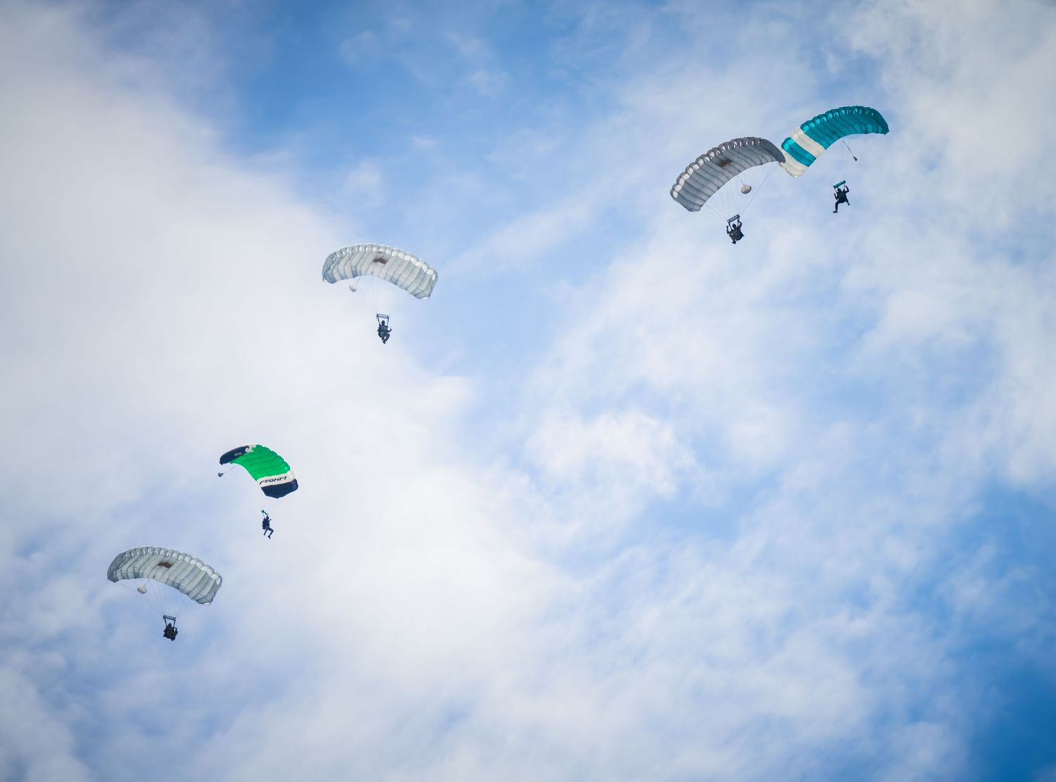 Skydivers with turquoise and white parachutes glide across a cloudy blue sky.
