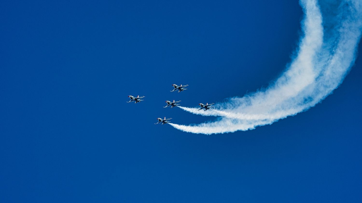 Aerobatic planes perform synchronized maneuvers leaving white smoke trails against bright blue sky.