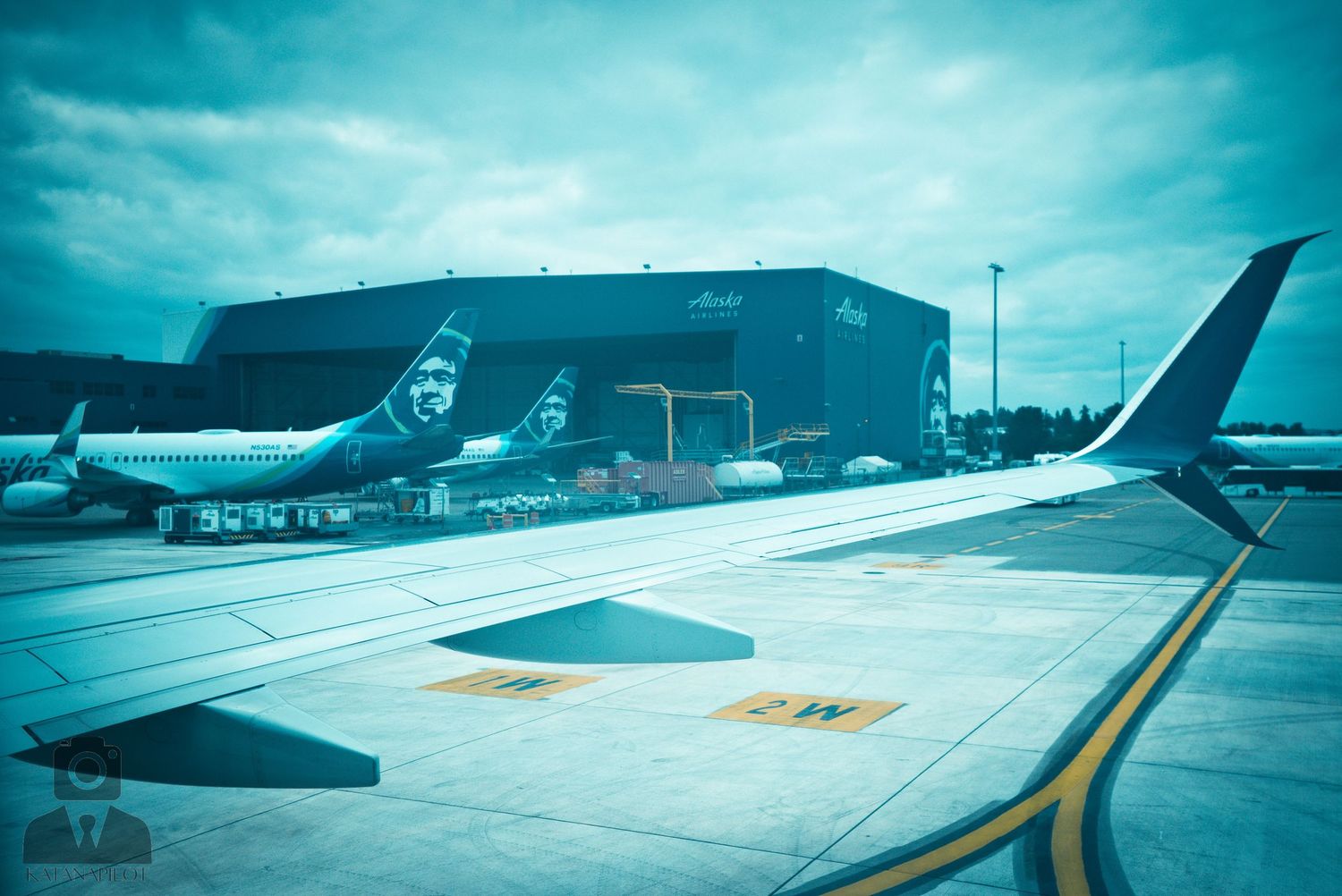 Aircraft on tarmac with teal-tinted sky and hangar buildings in background.