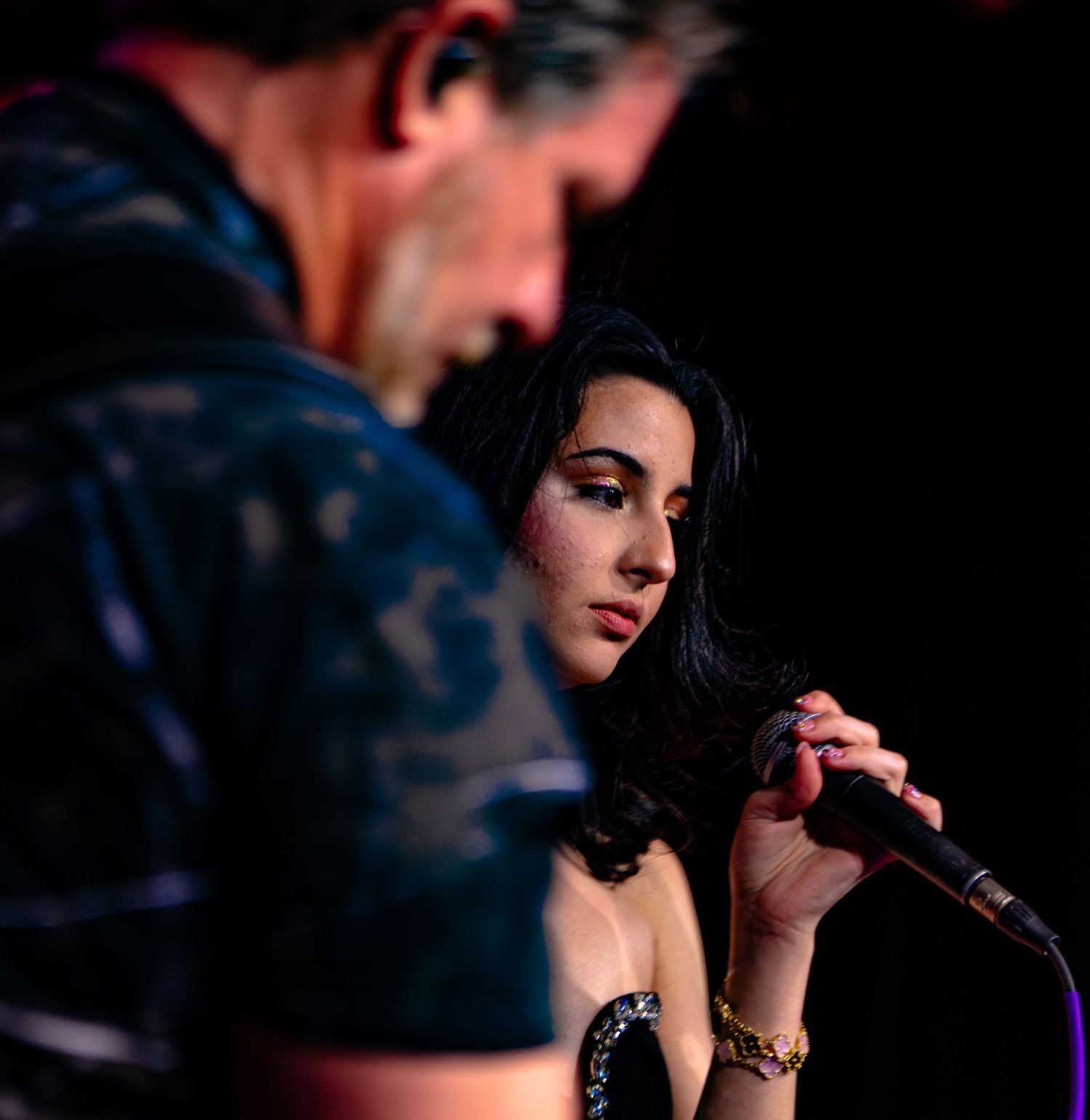 A young woman with dark hair sings into a microphone on stage beside an older man at a live music performance.