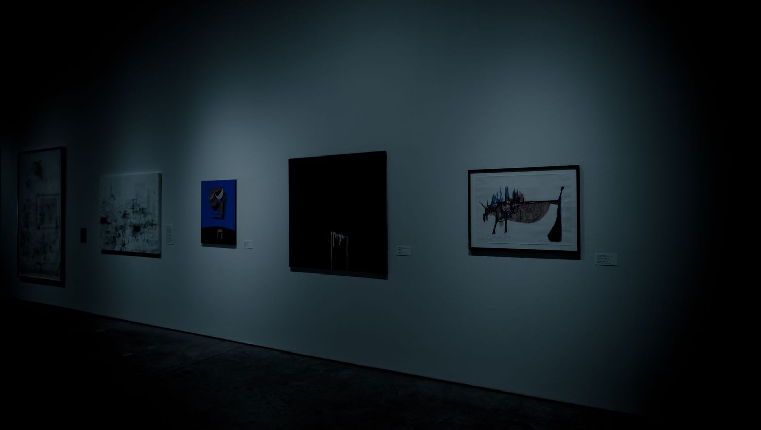 Dimly lit art gallery wall featuring mixed media artworks including blue and black abstract pieces.