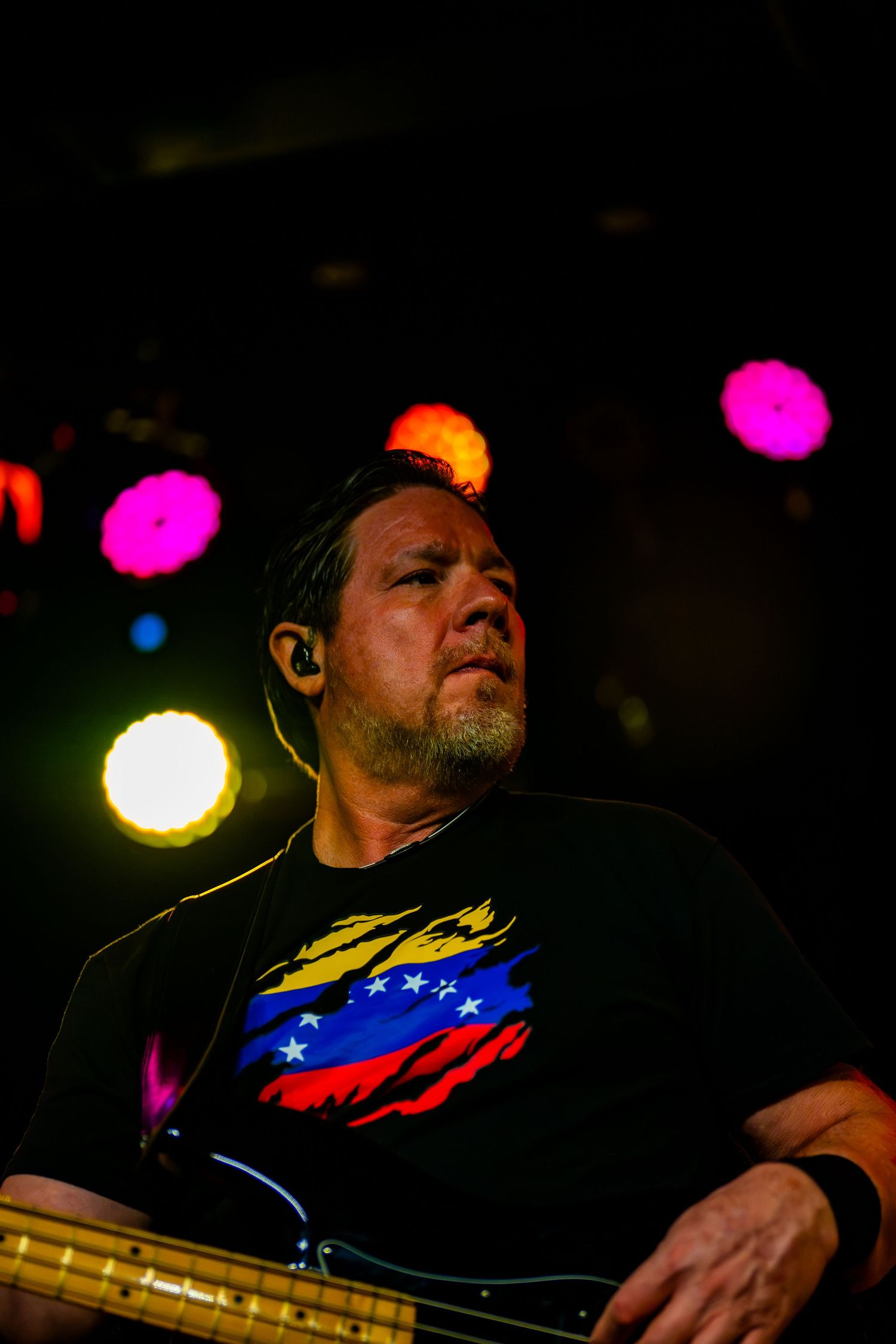 Male musician performing on stage with Venezuelan flag shirt, in-ear monitor, playing bass guitar under colorful concert lights.