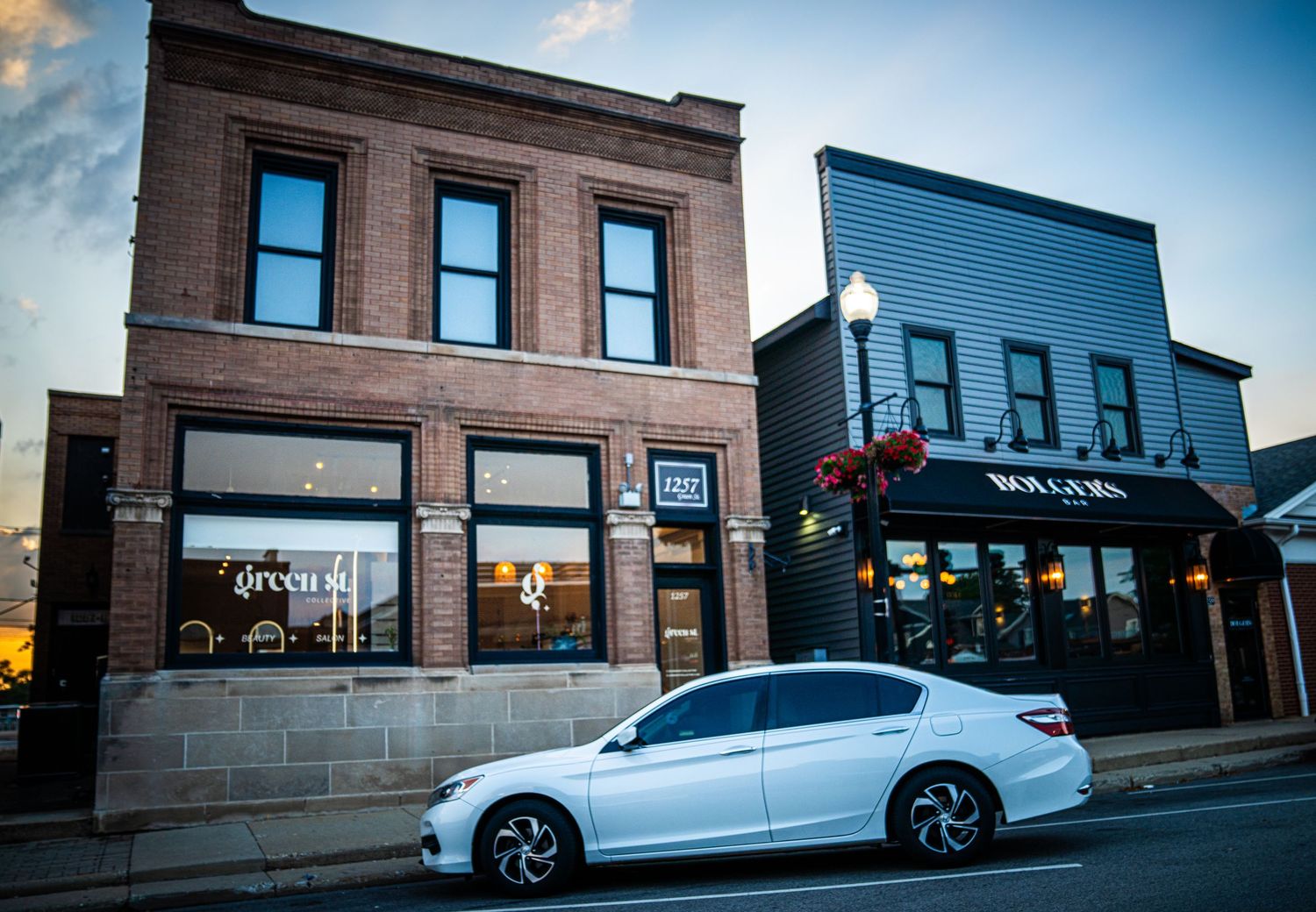 Two historic brick commercial buildings with large windows and parked car on street at dusk.