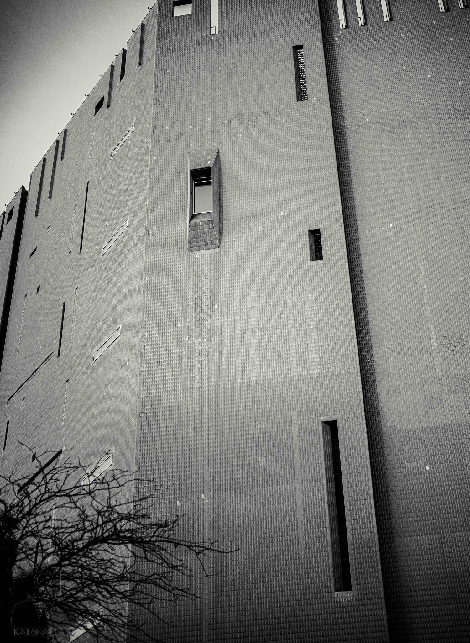 Black and white architectural photo of a tall brick building with narrow vertical windows and dark shadows.