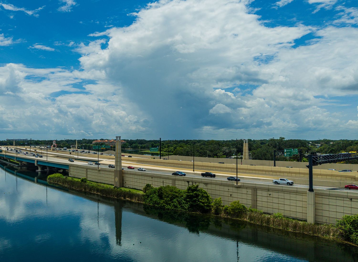Aerial view of a busy highway alongside a calm river under a blue sky with large white clouds and lush green trees.