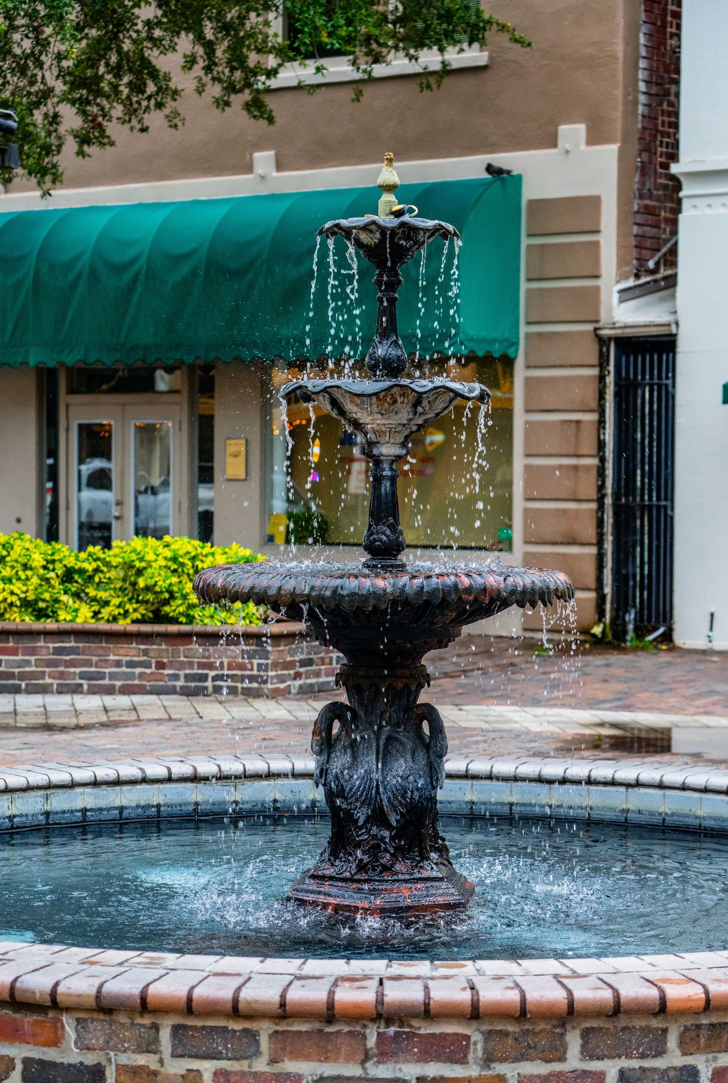 Ornate black fountain spraying water in a brick-lined pool with green awning in background.