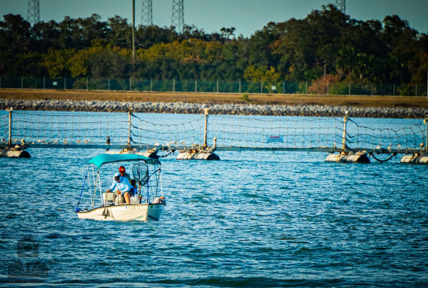 Row of sailboats and watercraft line up along a waterway with trees in the background.