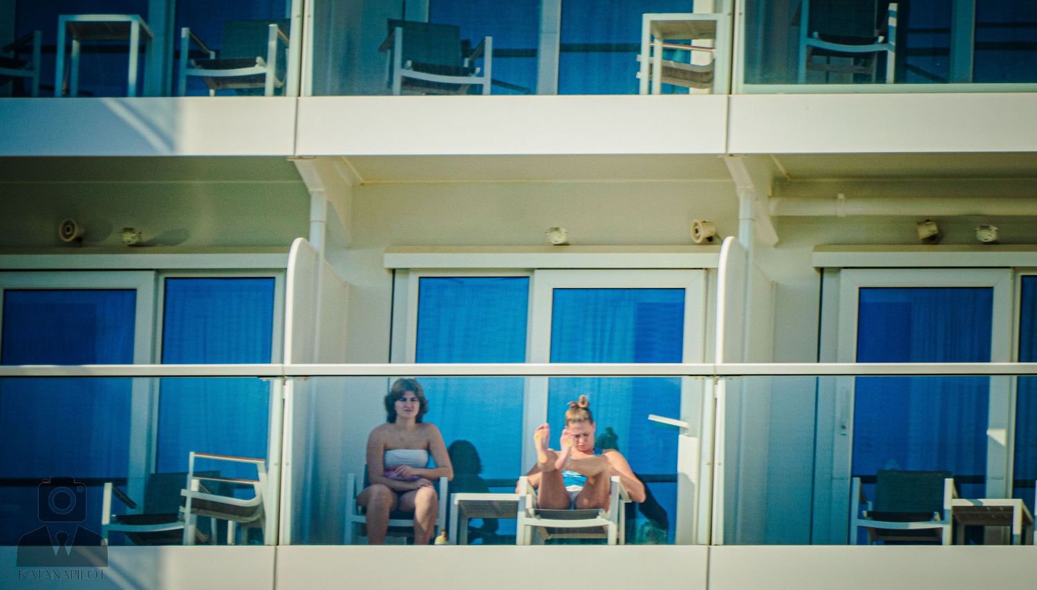 Two people relax on balcony chairs of a cruise ship with blue glass windows and white exterior.