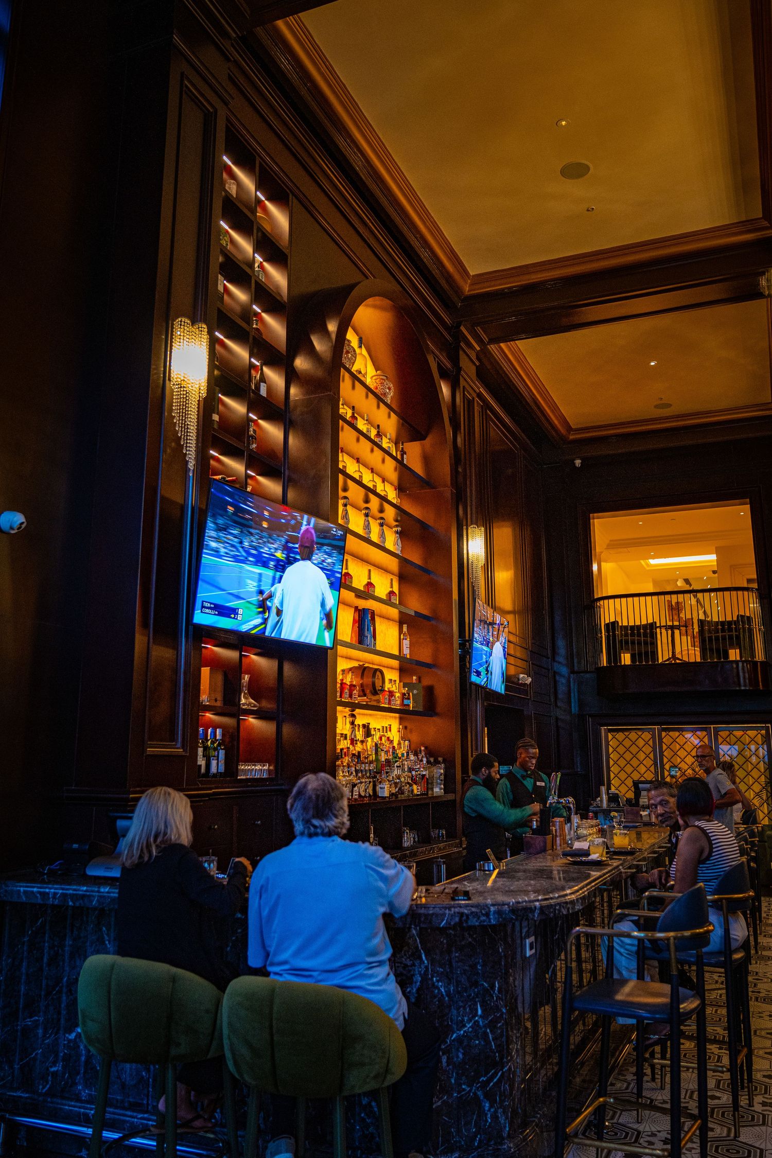 Elegant upscale bar with dark wood shelving, glowing TV screens, and patrons seated at a marble counter.