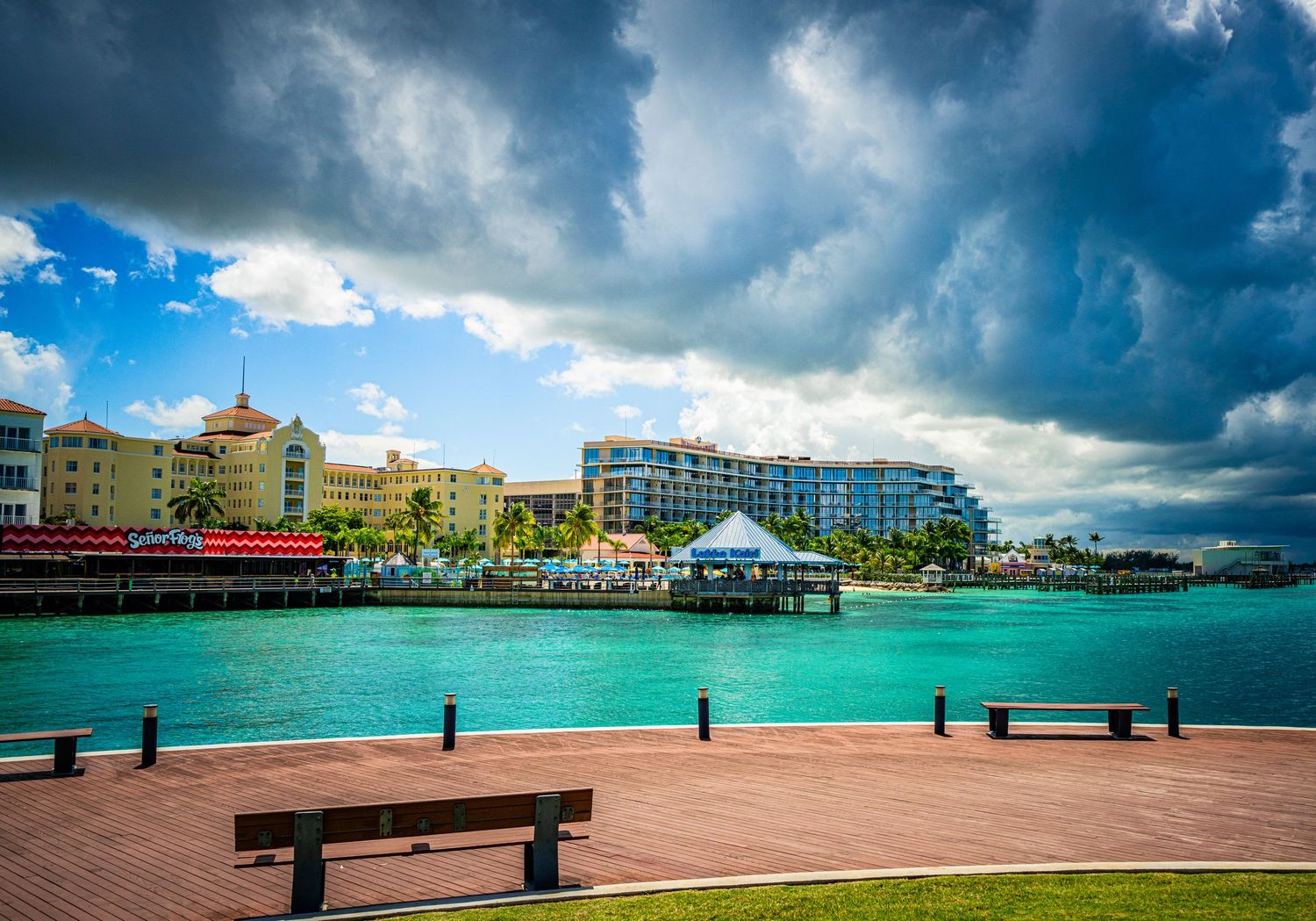 Turquoise waterfront with colorful buildings, a pier, and dramatic storm clouds over a tropical harbor.