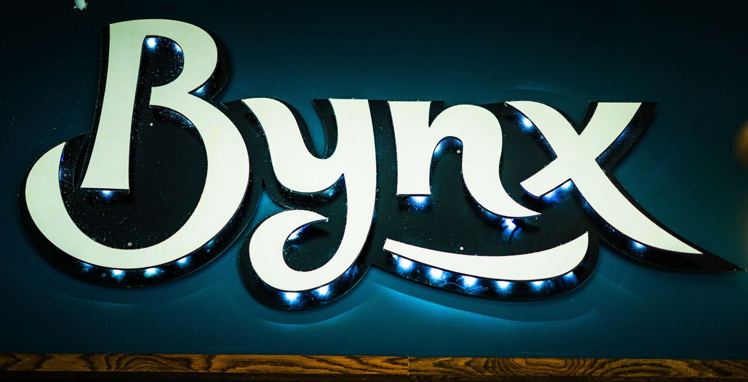 Illuminated Bynx logo sign with LED lights on a teal wall, featuring elegant script lettering.