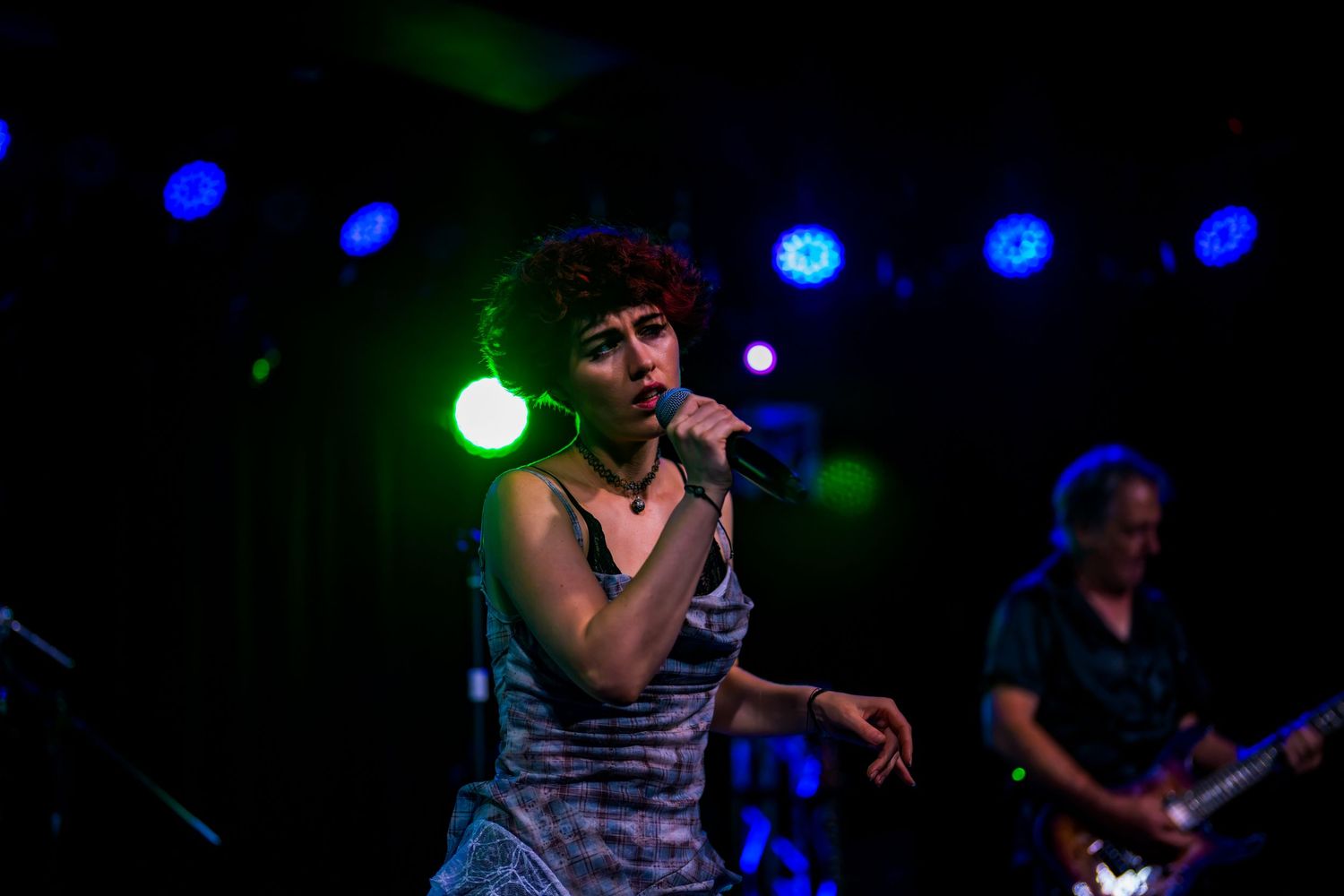 Female vocalist with curly hair sings into microphone on dark stage with blue concert lighting.