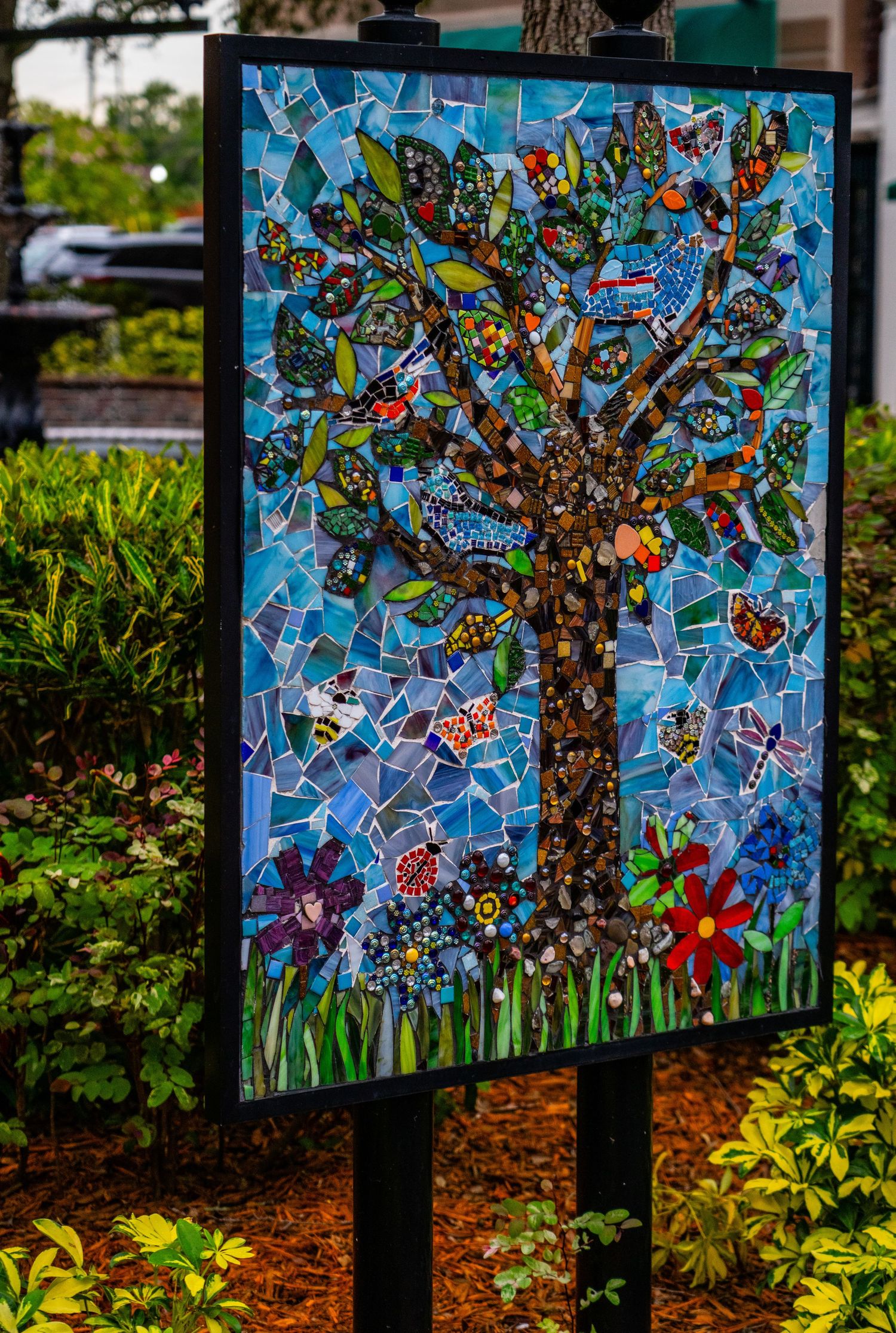 Vibrant mosaic art piece featuring a colorful tree design against a blue background displayed outdoors.