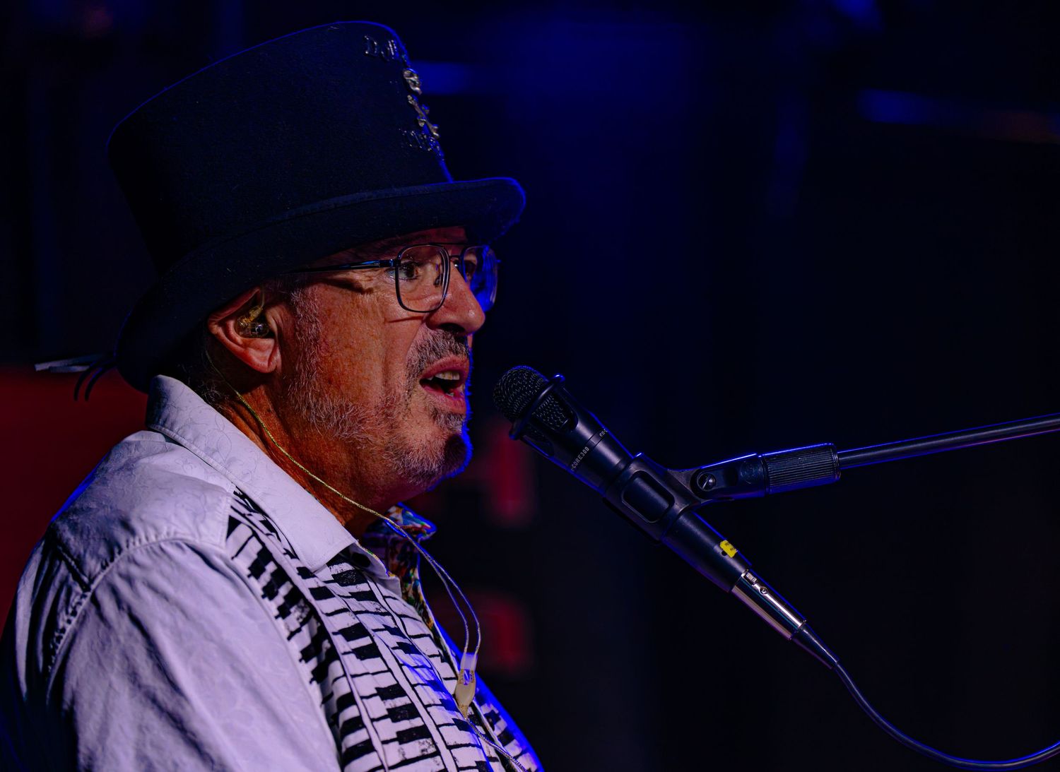 Man in fedora hat singing at microphone on stage during live music performance.