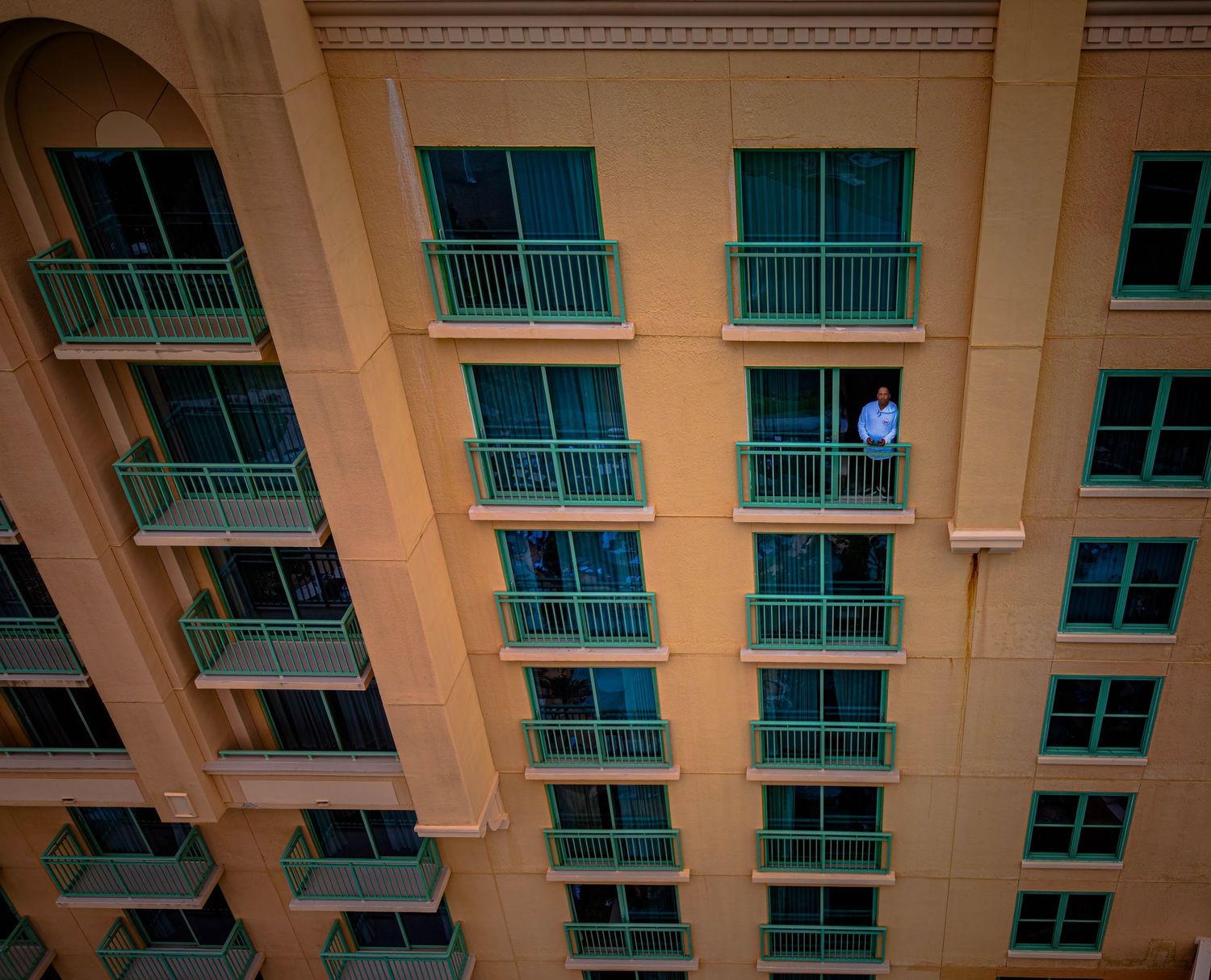 Person standing on balcony of large yellow hotel building with green-trimmed windows and multiple floors.