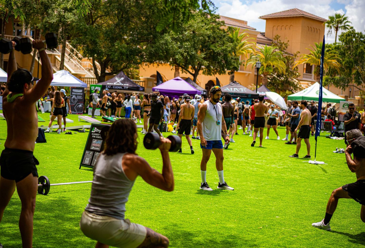 Outdoor fitness event on green turf with people exercising, tents, and palm trees on a sunny day.