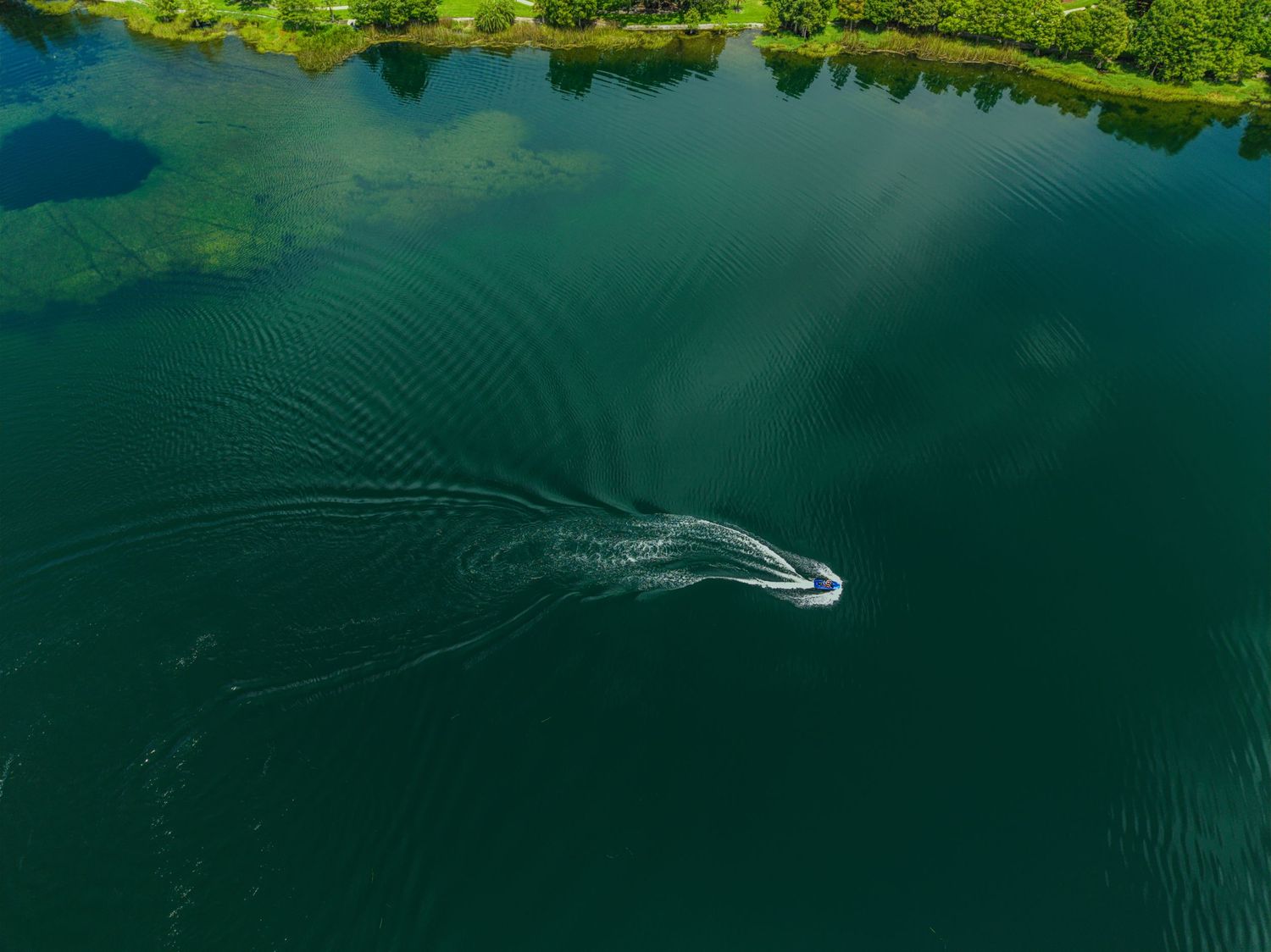 Aerial drone view of a speedboat creating a dramatic wake pattern on a deep green lake surrounded by trees.