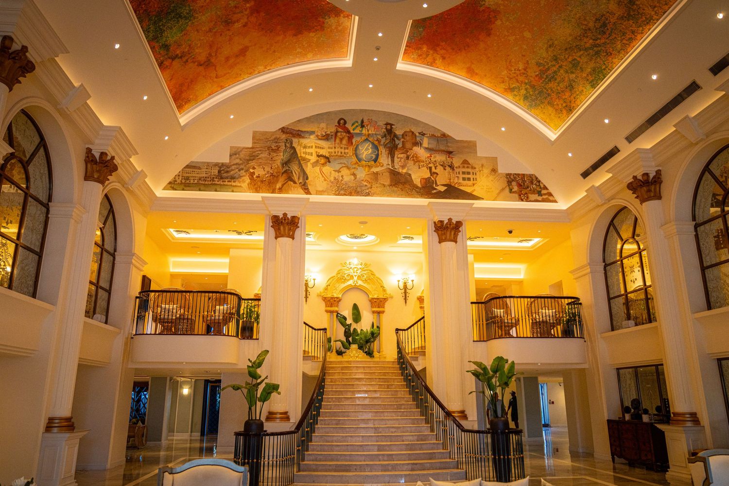 Elegant hotel lobby with grand staircase, ornate ceiling mural, white columns, and warm golden lighting.
