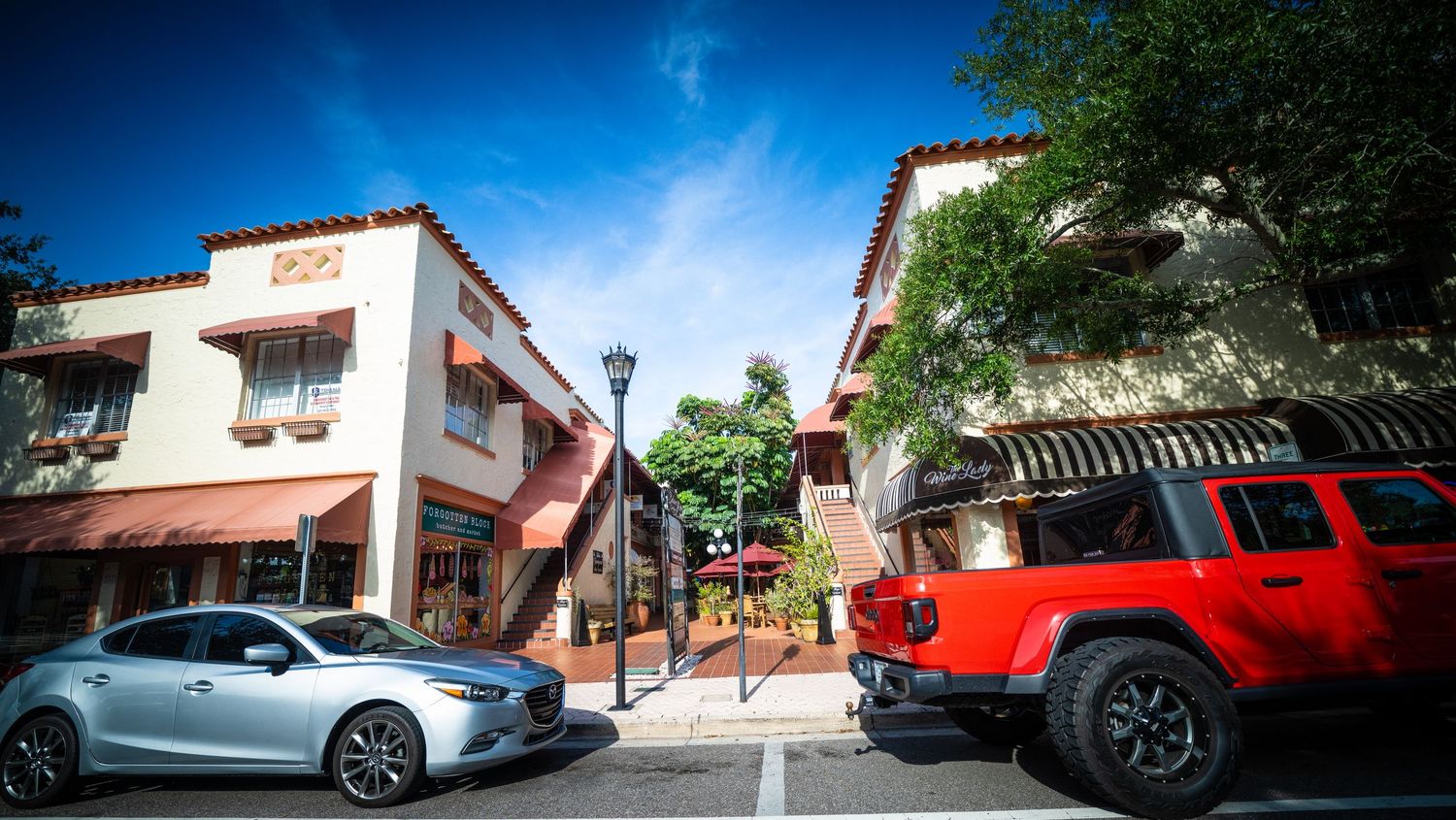 Mediterranean-style shopping plaza with red and silver vehicles parked in front under blue sky.