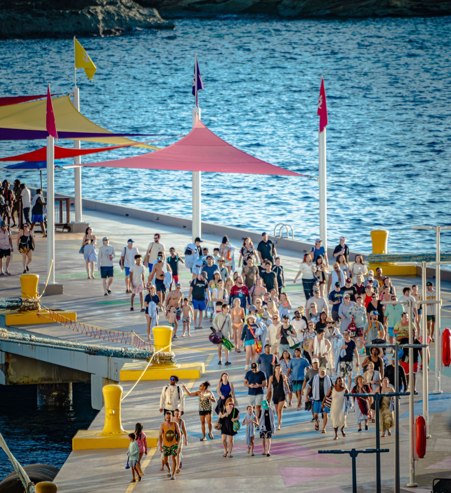 Crowds of beachgoers walk along a waterfront promenade with colorful canopies and flags on a sunny summer day.