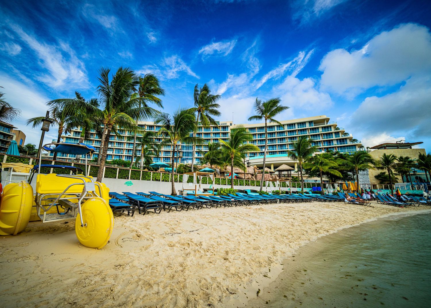 Tropical beachfront hotel with blue lounge chairs, palm trees, and white sandy beach under a vibrant blue sky.