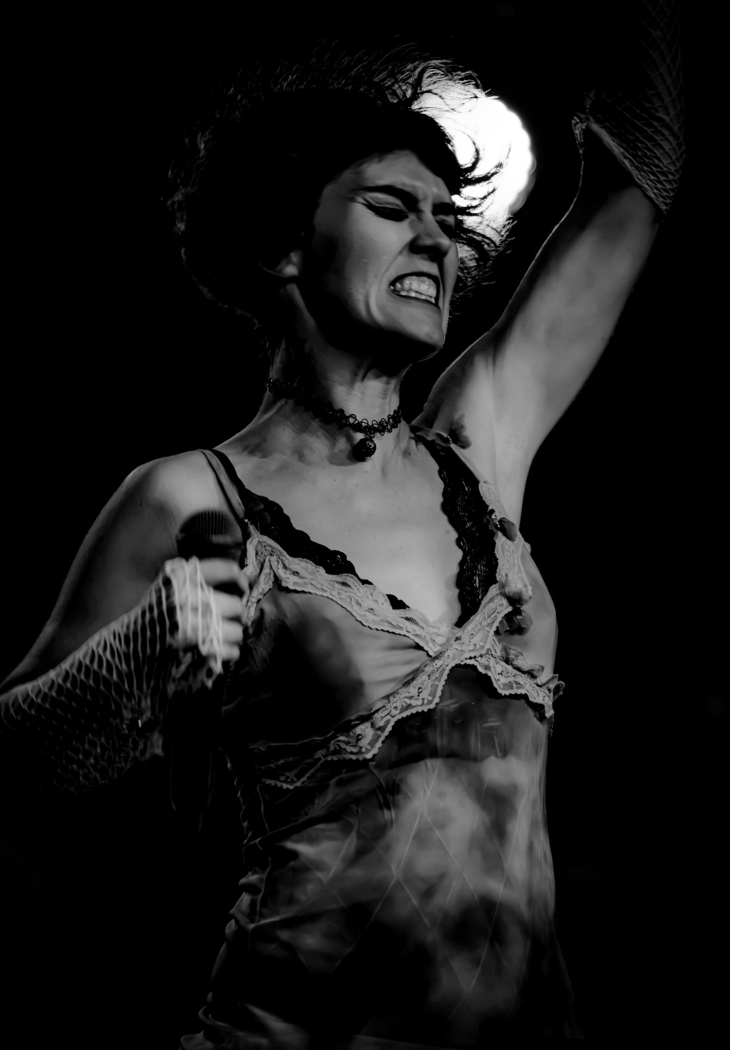Black and white dramatic photo of a woman performing on stage in lingerie with arm raised and intense expression.
