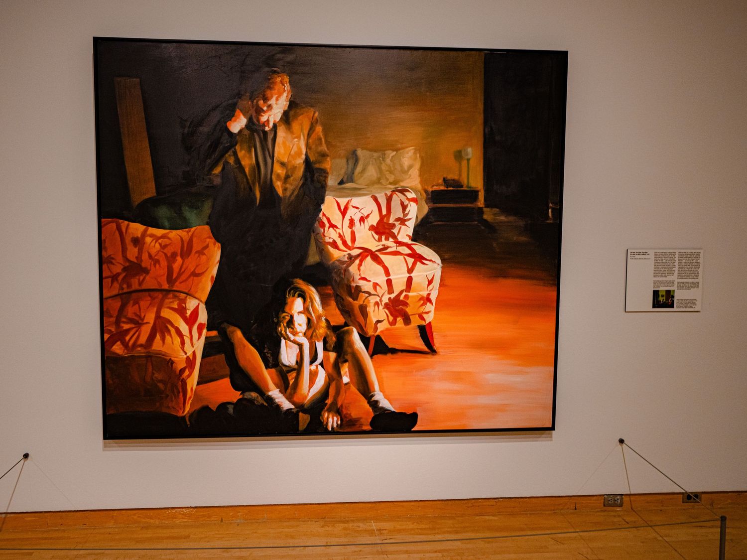 Large oil painting in gallery showing figures with orange armchairs in warm tones, displayed on white museum wall.