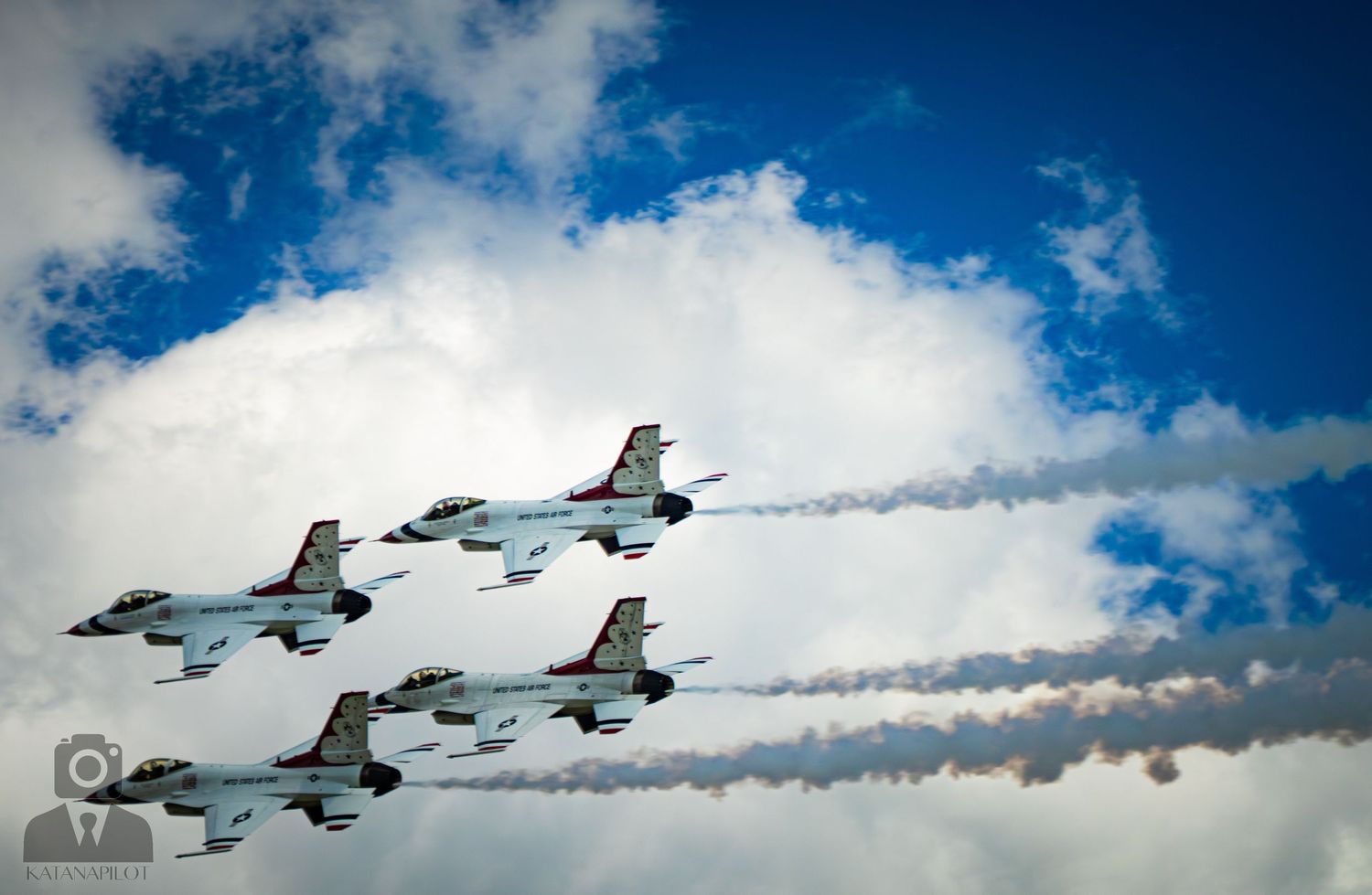Four USAF Thunderbirds fighter jets fly in tight diamond formation leaving white smoke trails against a blue cloudy sky.