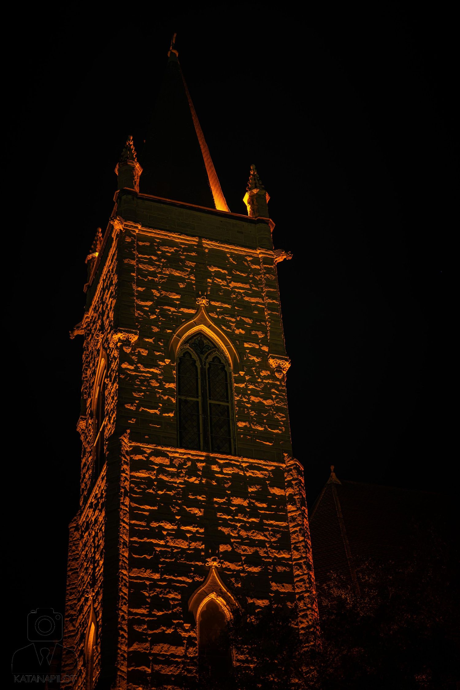 Gothic stone church steeple illuminated by warm orange light against a dark night sky.