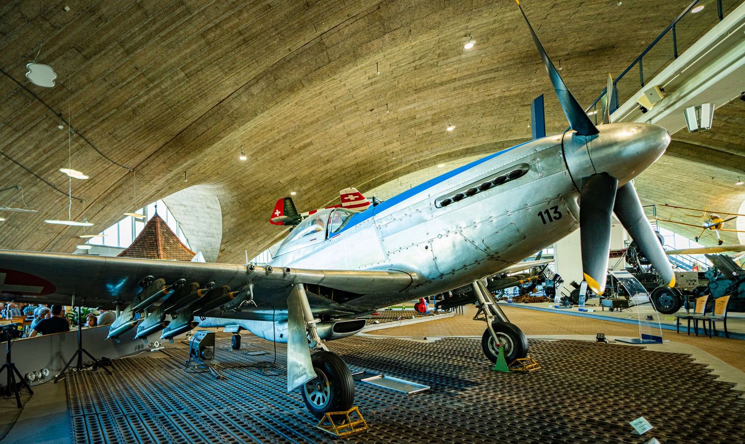 Vintage silver propeller fighter plane with blue accents on display inside a curved-roof aviation museum hangar.