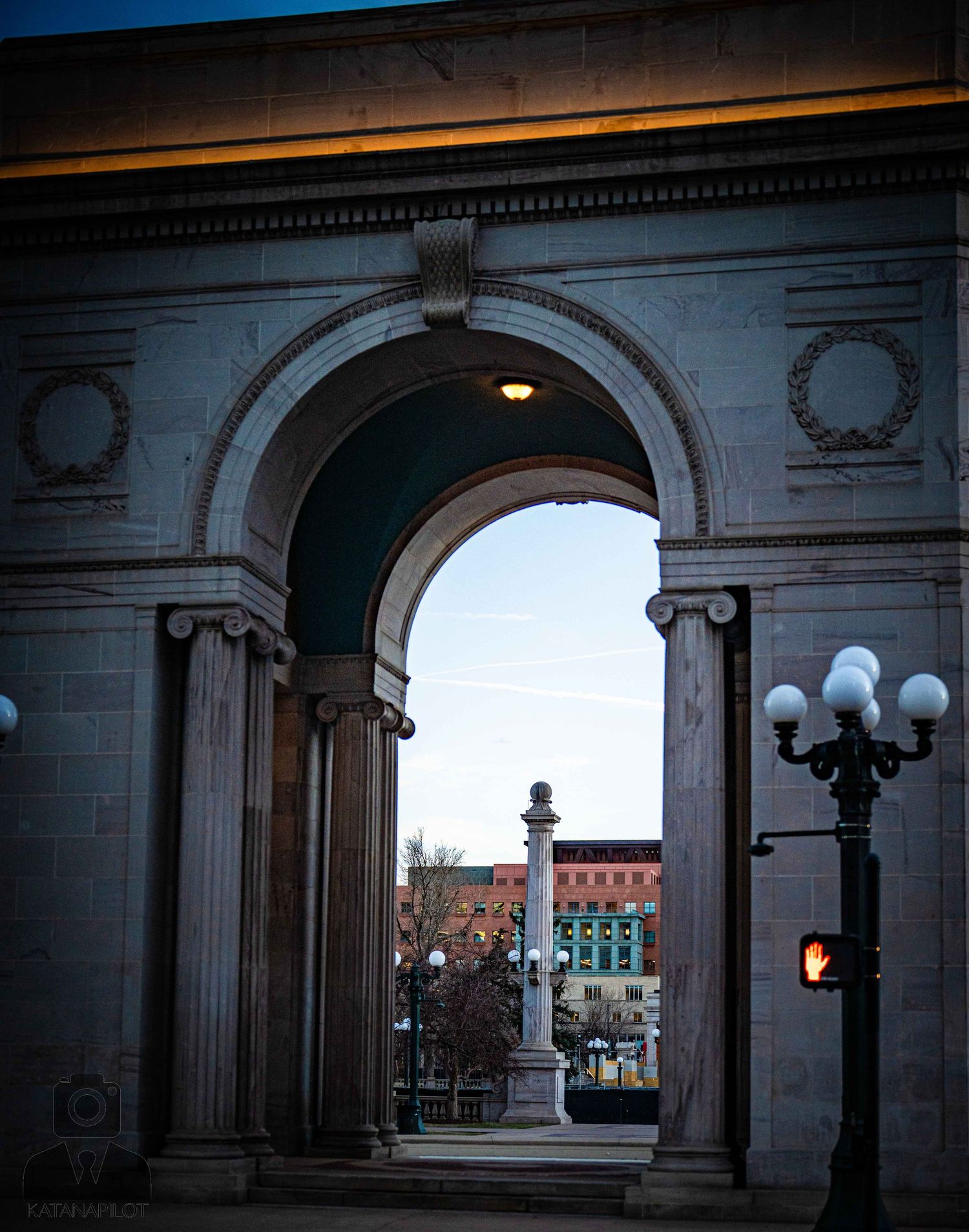 Architectural arches frame a view of ornate lamp posts and buildings in a historic downtown area at dusk.